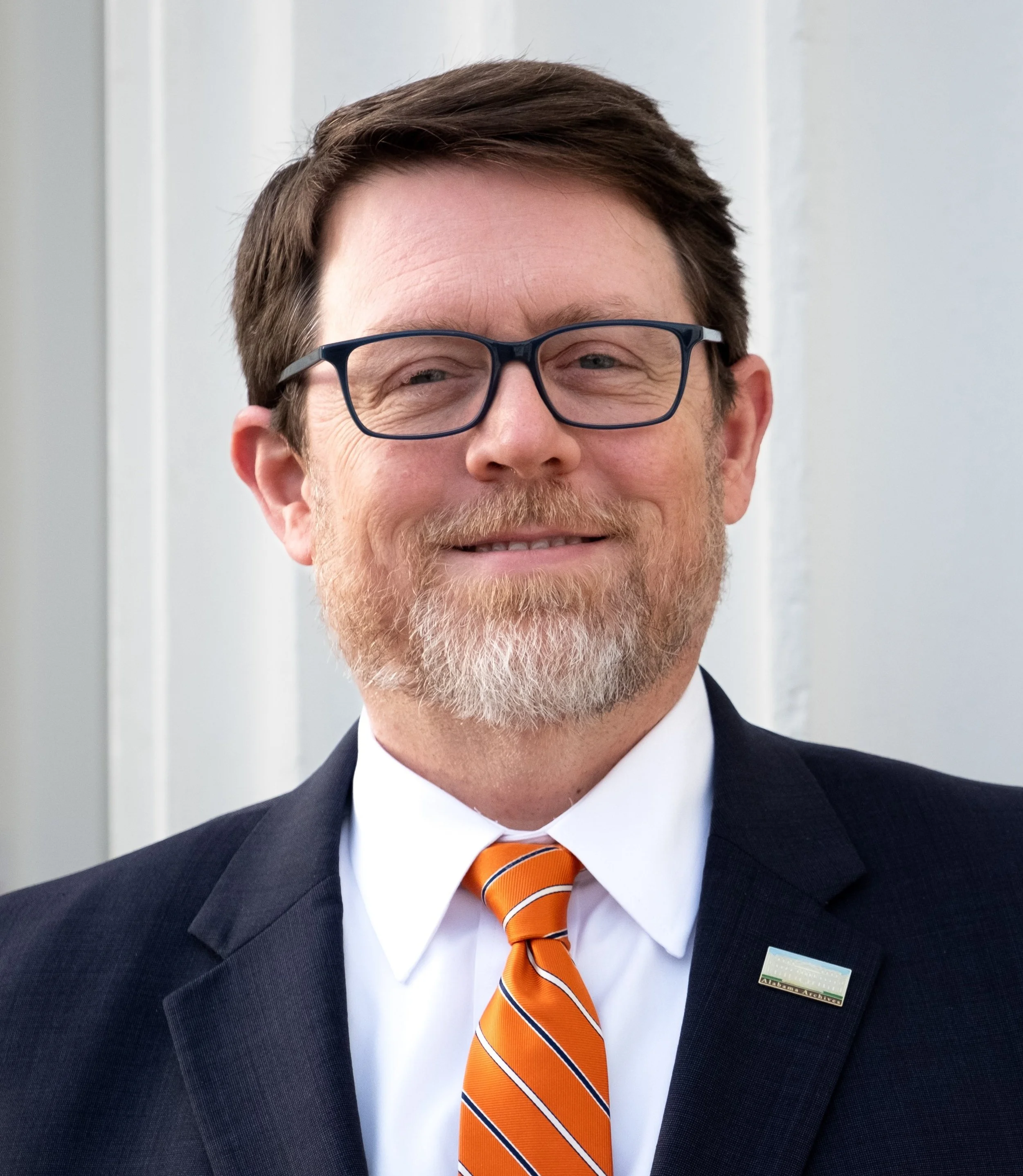 Steve Murray with glasses, a beard, and mustache, dressed in a suit, white shirt, and an orange striped tie, smiling.