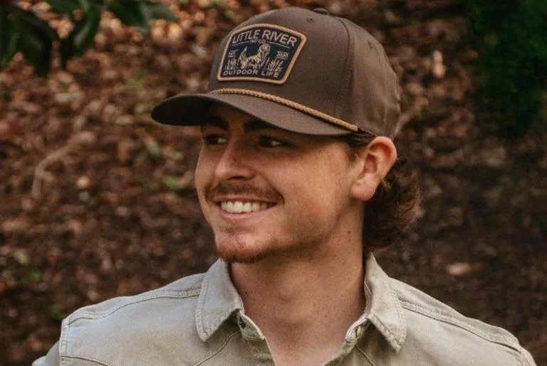 Myles Morgan smiling outdoors, wearing a gray cap and a light-colored button-up shirt, with a background of brown leaves.