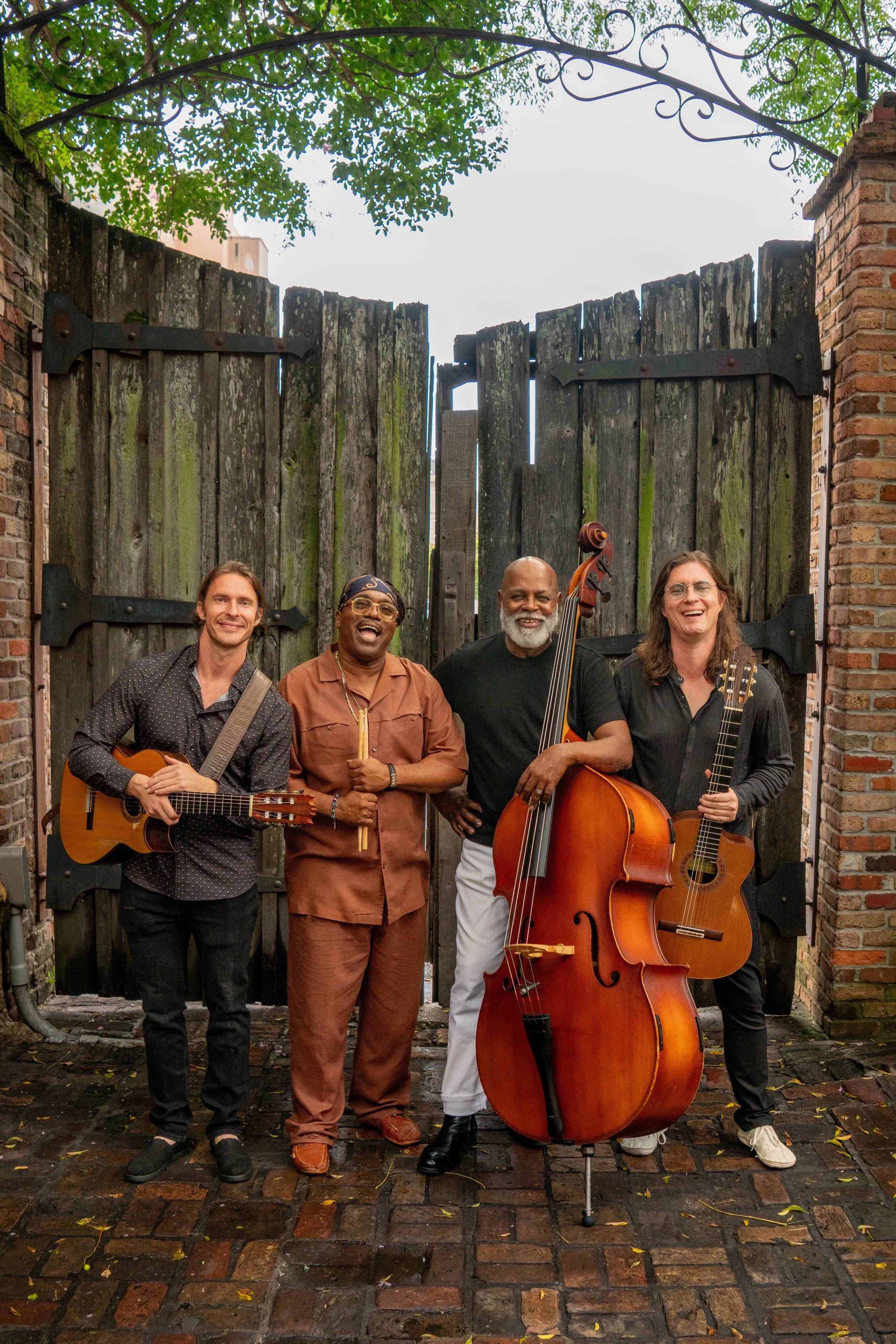 Roman Street standing outdoors, holding instruments: two guitars, a violin, and a double bass, smiling at the camera.