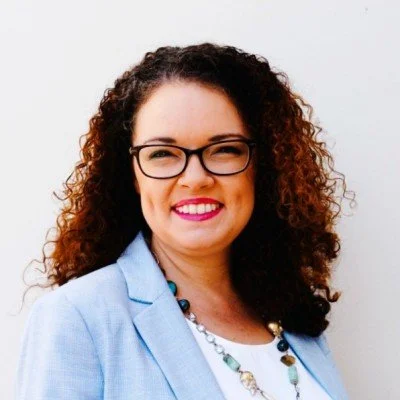 Kristin Hellmich with curly hair wearing glasses, a light blue blazer, and a necklace, smiling at the camera.