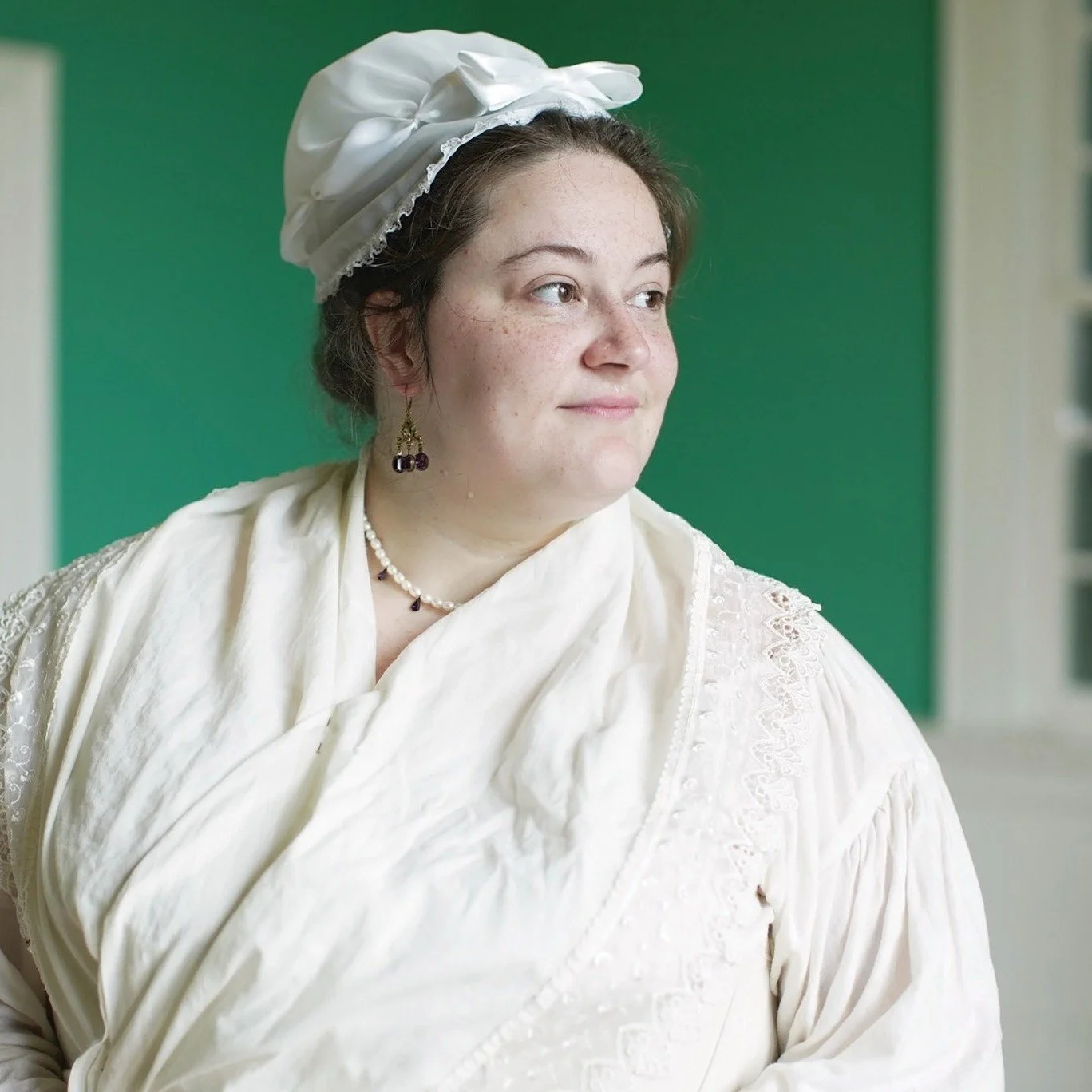 A young woman with short brown hair, wearing a white bonnet and vintage-style cream-colored dress with lace details, standing indoors against a green background.