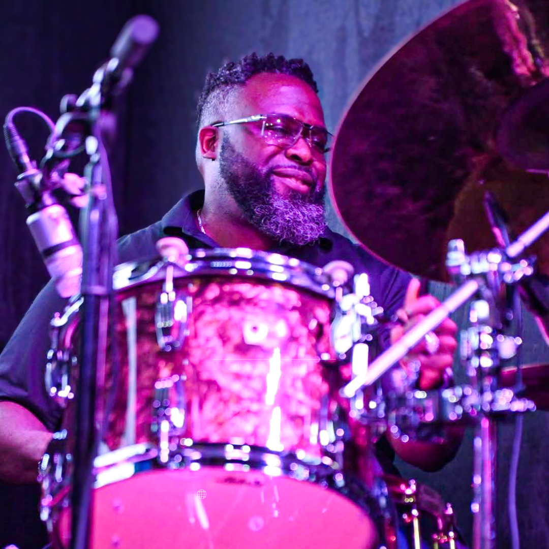 PJ Spraggins playing drums on stage, surrounded by purple and pink lighting.