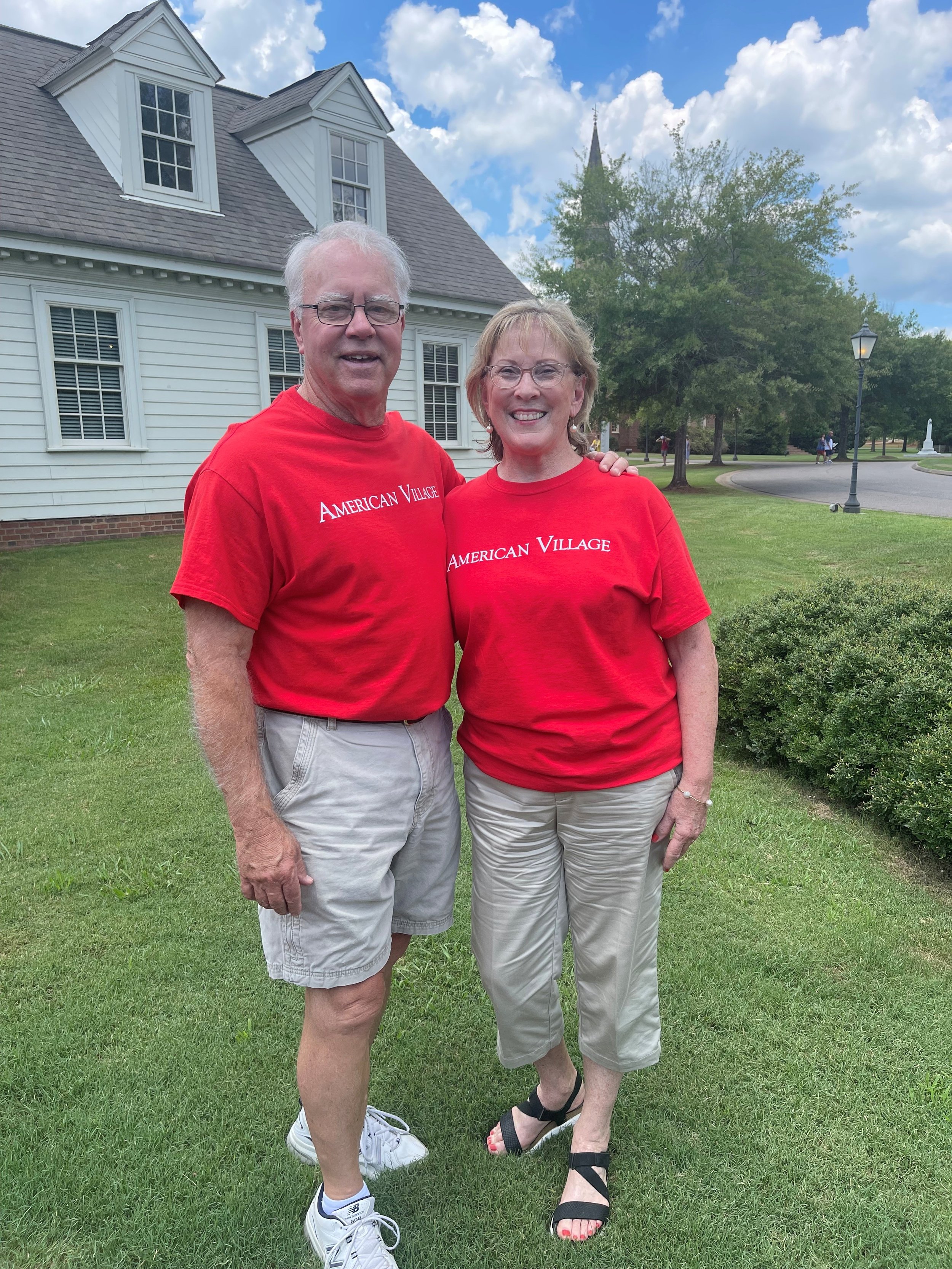 A man and woman standing outside on a grassy area, both wearing red t-shirts that say 'American Village,' smiling and posing for a photo. Behind them are trees, a white house with multiple windows, and a small church steeple.
