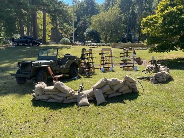 An outdoor scene with a vintage military Jeep, sandbags arranged as a barrier, and various wooden and concrete barriers in a grassy area surrounded by trees.