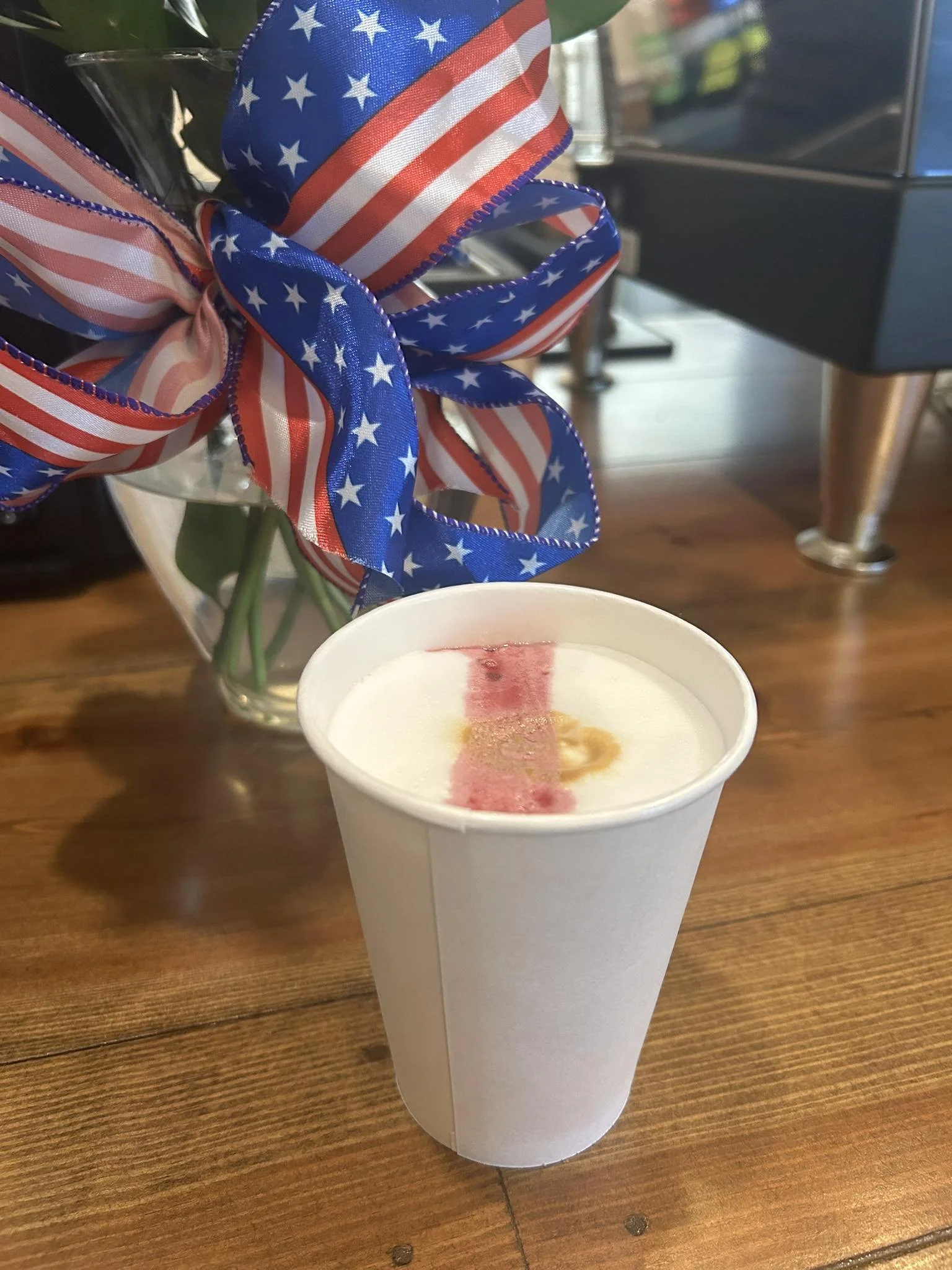 A white paper cup filled with a beverage, possibly coffee, with some pink and yellowish markings on top, placed on a wooden table. In the background, there is a clear glass vase with pink, white, and blue ribbons with stars and stripes, tied into a bow, and some green leaves visible inside the vase.