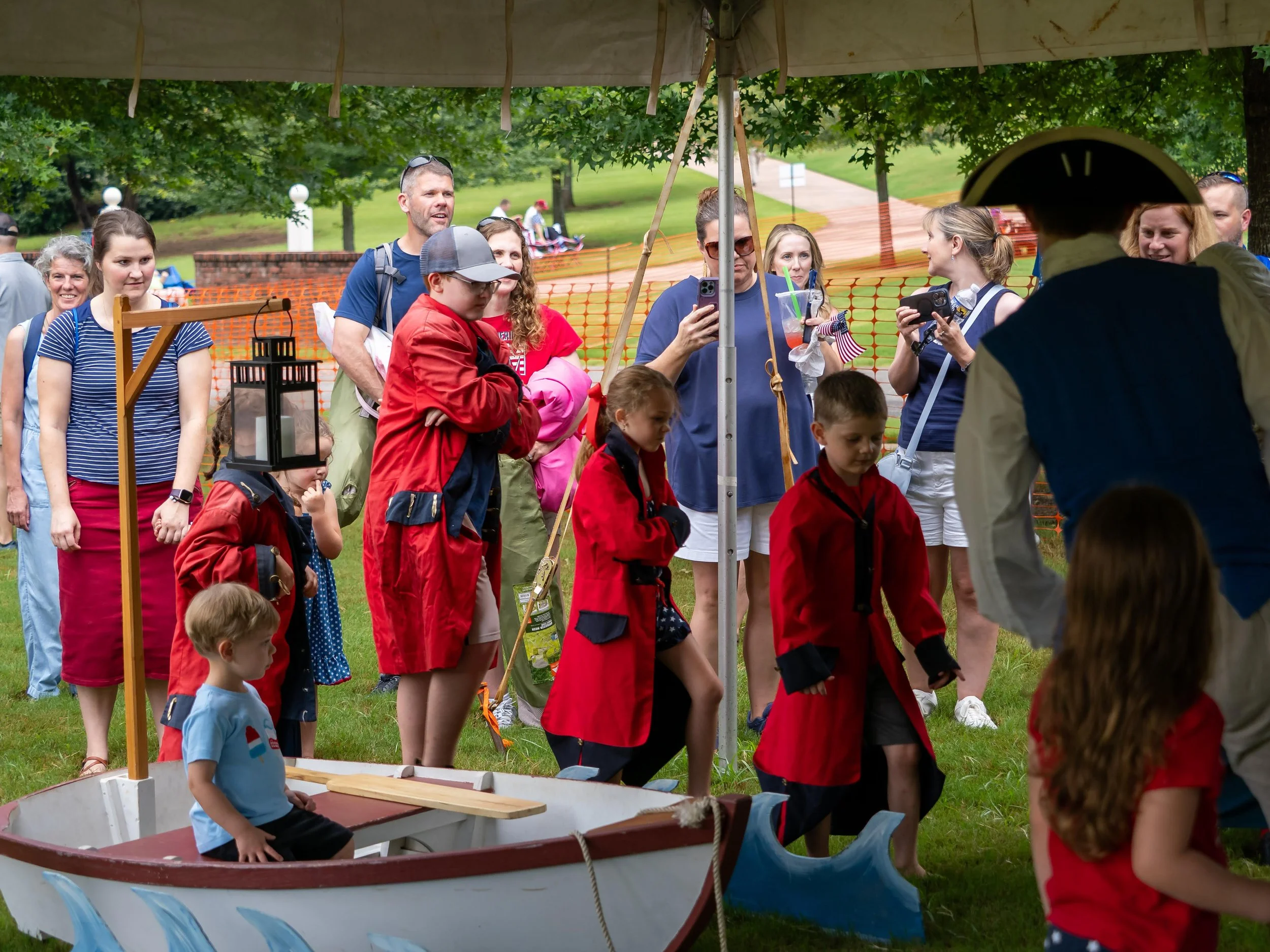 Children and adults gathered around a reenactor in colonial attire at an outdoor event, with a small boat and a white tent.