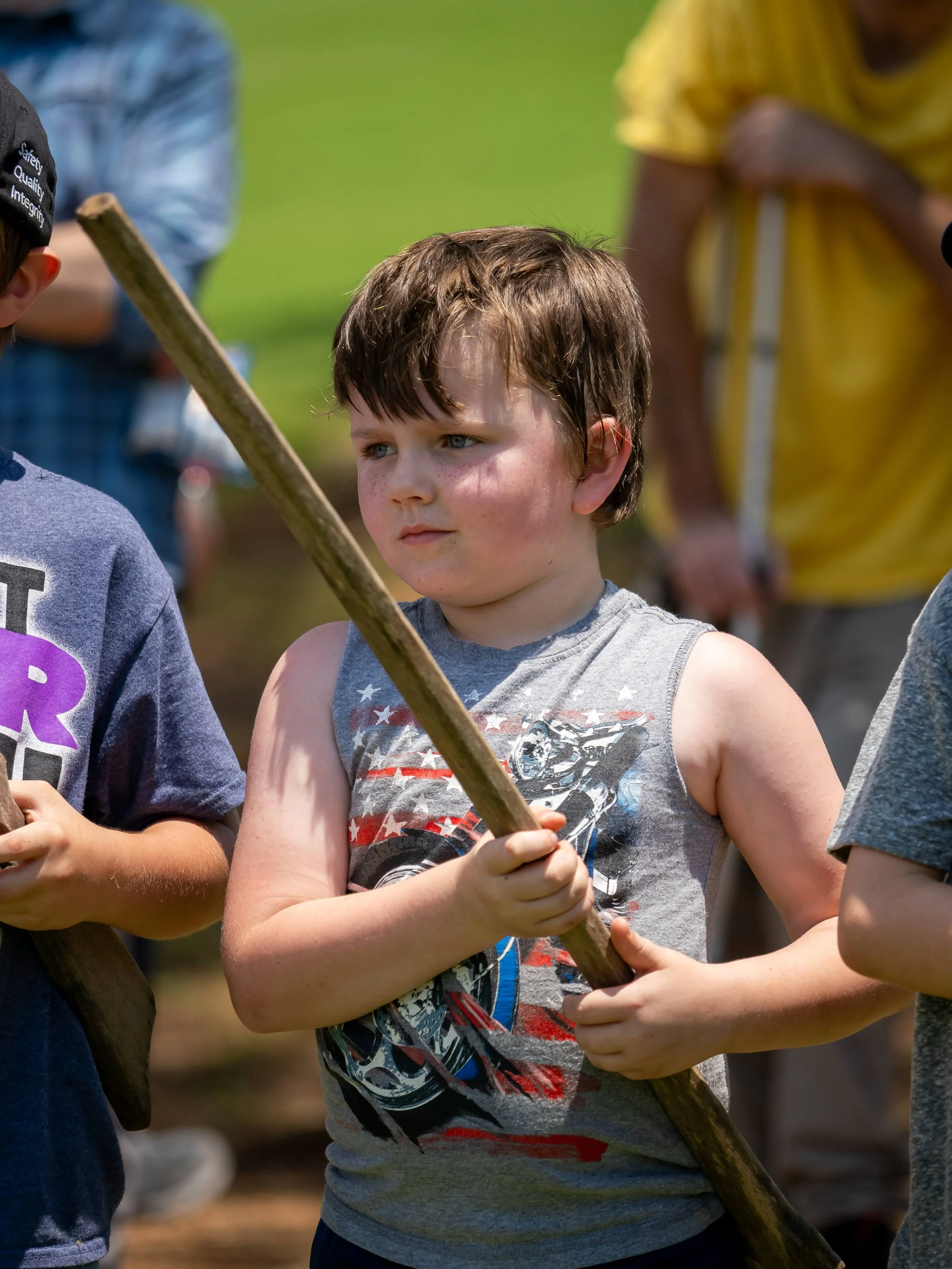 A young boy with brown hair and fair skin holding a stick, standing outdoors during daytime. He is wearing a sleeveless gray t-shirt with a graphic and appears to be focused. Other children and adults are visible in the background.