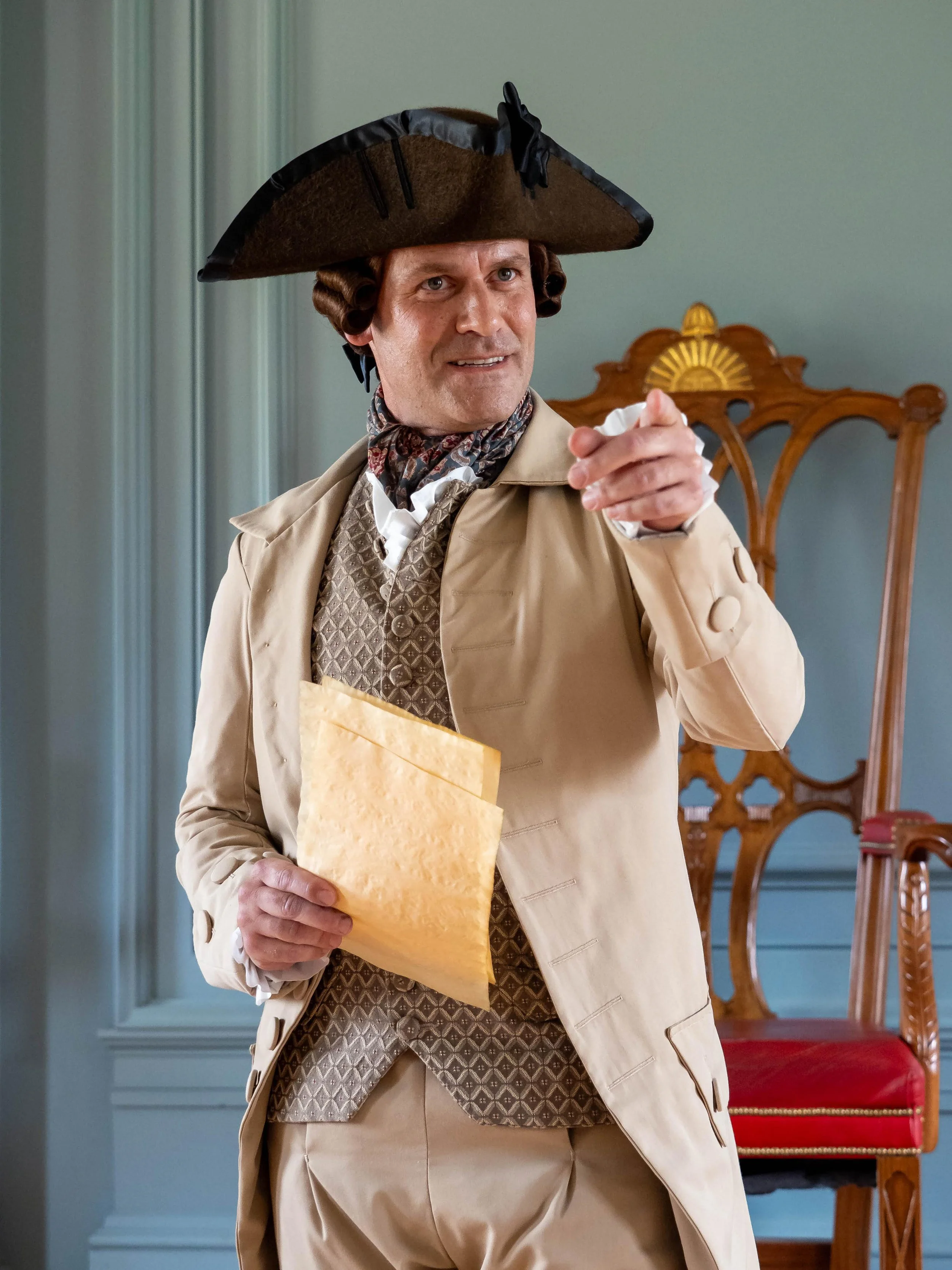A man dressed as a historical figure from the 18th century, wearing a tricorn hat, a beige coat, and patterned waistcoat, holding a yellow envelope and pointing with his finger.