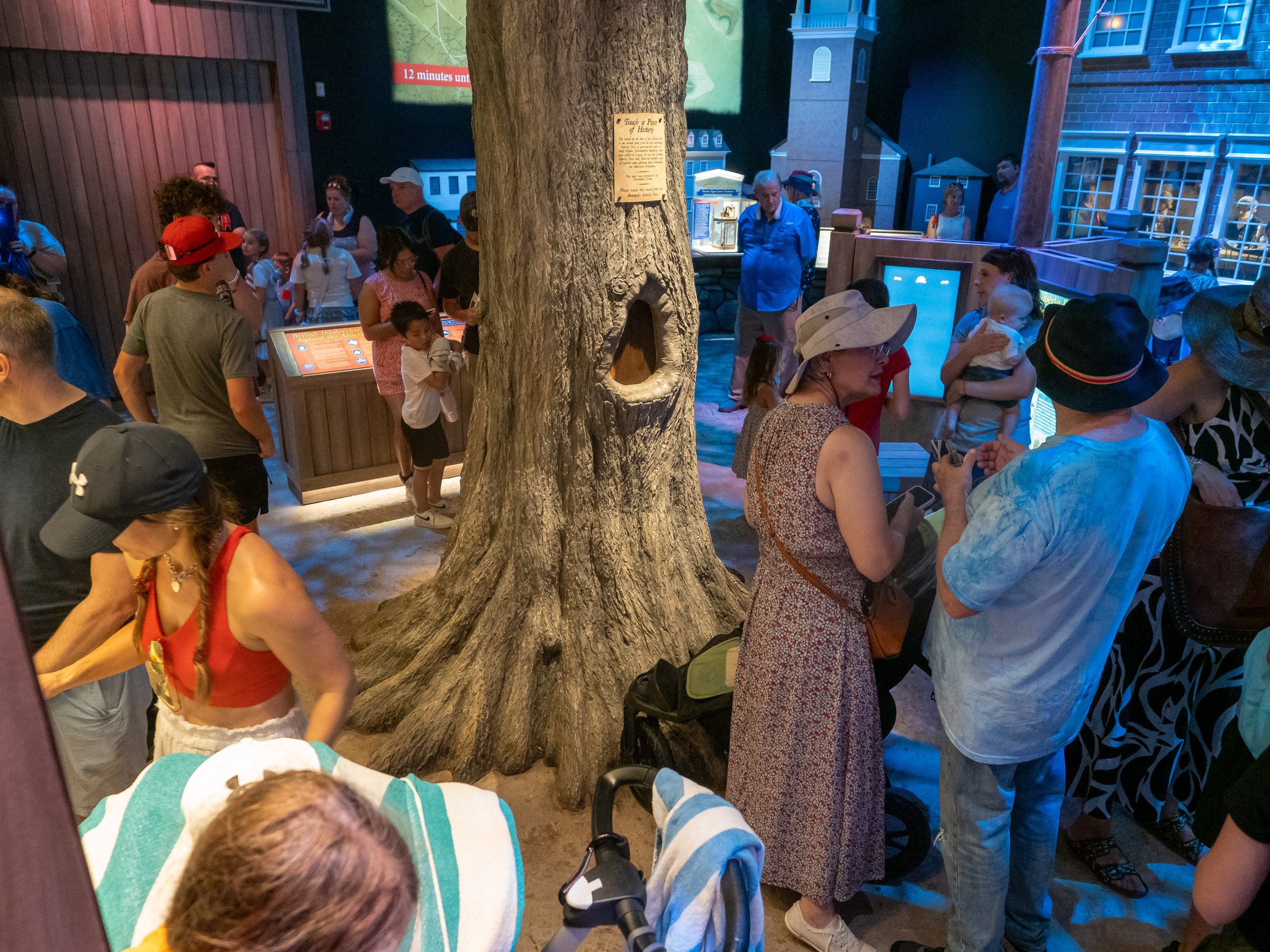 A crowded indoor museum or exhibit space with people of all ages gathered around a large tree trunk with a birdhouse. There are informational displays, a small model building, and a visible map on the wall in the background. Some people are engaged in conversations, others look at the displays or take photos.