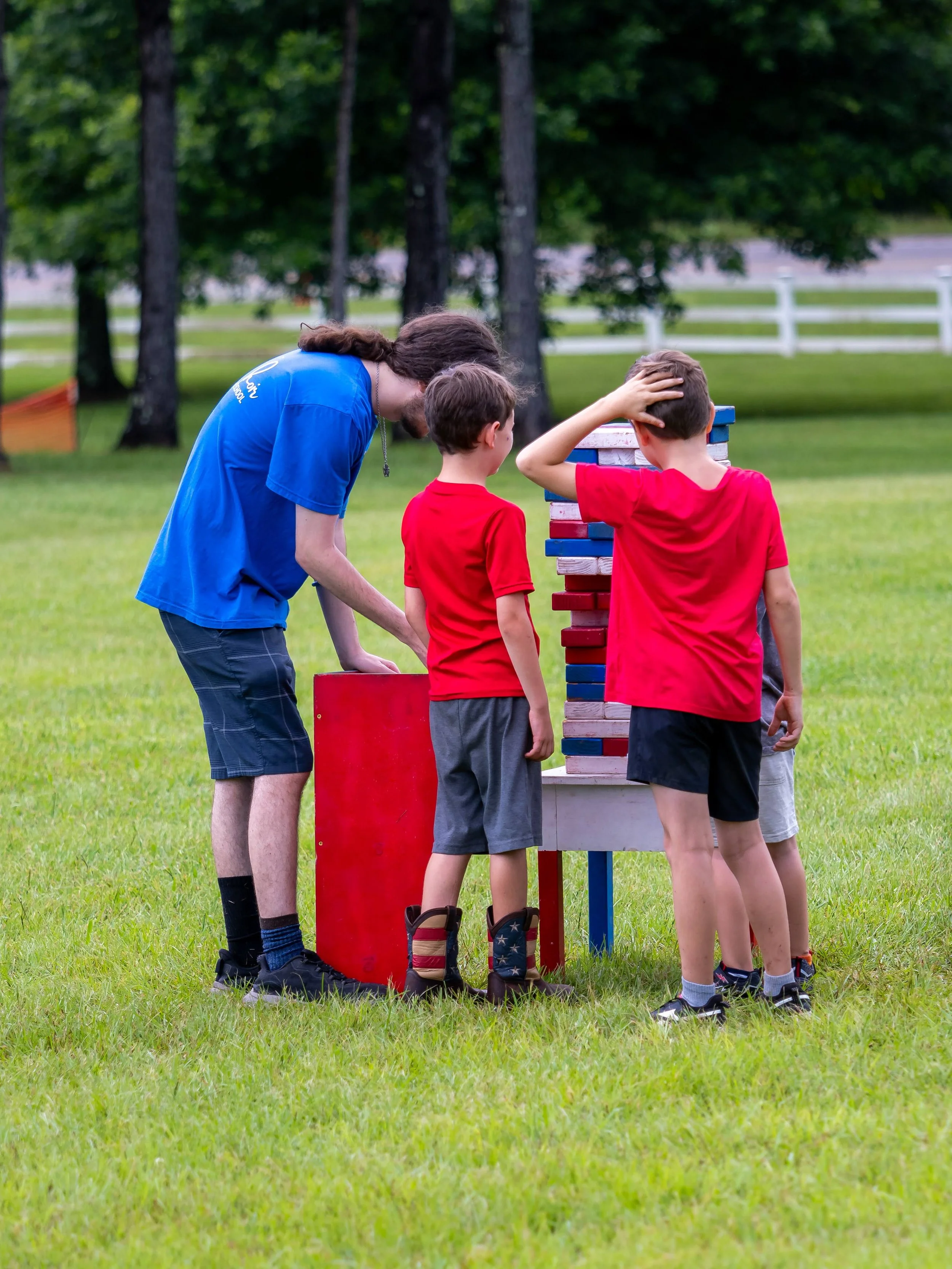 A man and three boys playing giant Jenga outdoors on a grassy field, with trees and a white fence in the background.