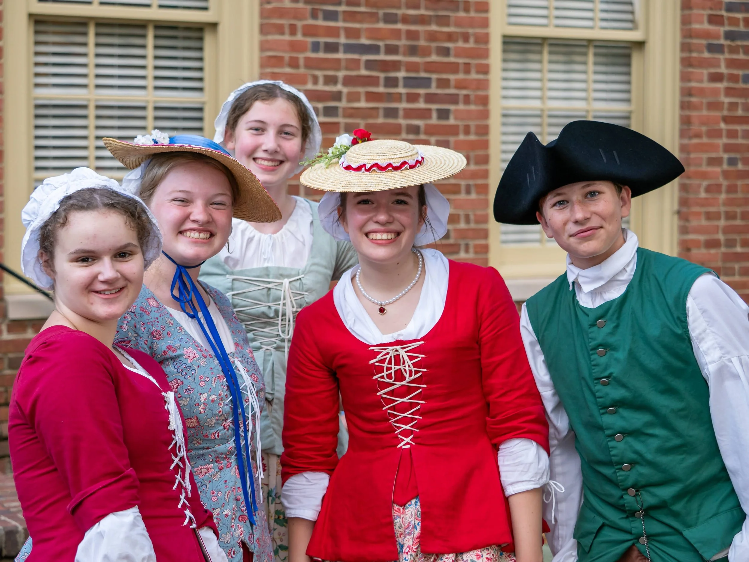 Group of young people dressed in historical costumes, smiling outdoors in front of a brick building.
