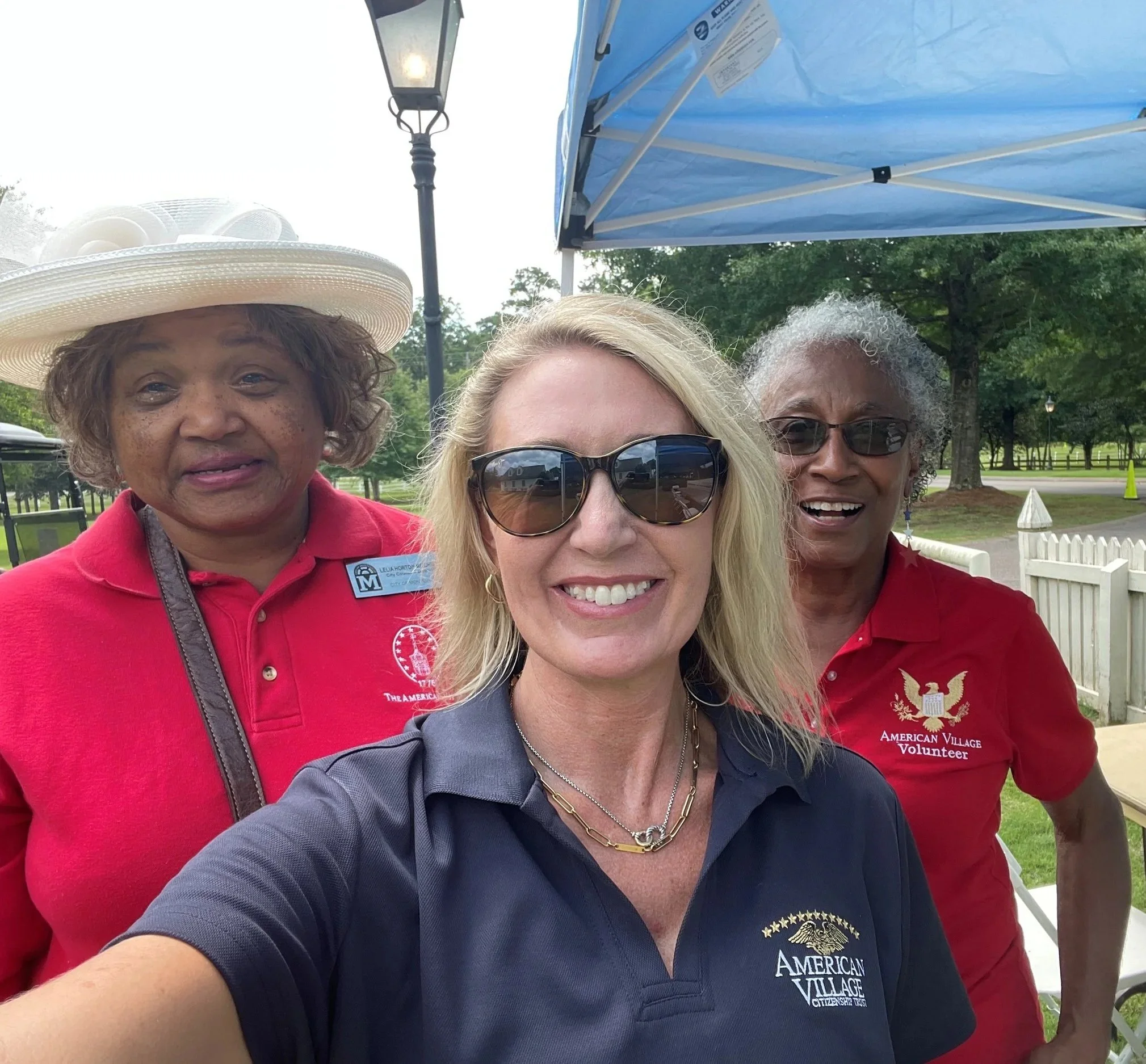 Three women smiling outdoors at a park or community event. The woman in the front is wearing sunglasses and a navy blue shirt with an 'American Village' logo. The woman on the left is wearing a red polo shirt with a name tag and a large hat. The woman on the right is wearing a red polo shirt with an 'American Village Volunteer' logo. There is a blue canopy tent, a street lamp, trees, and a white fence in the background.