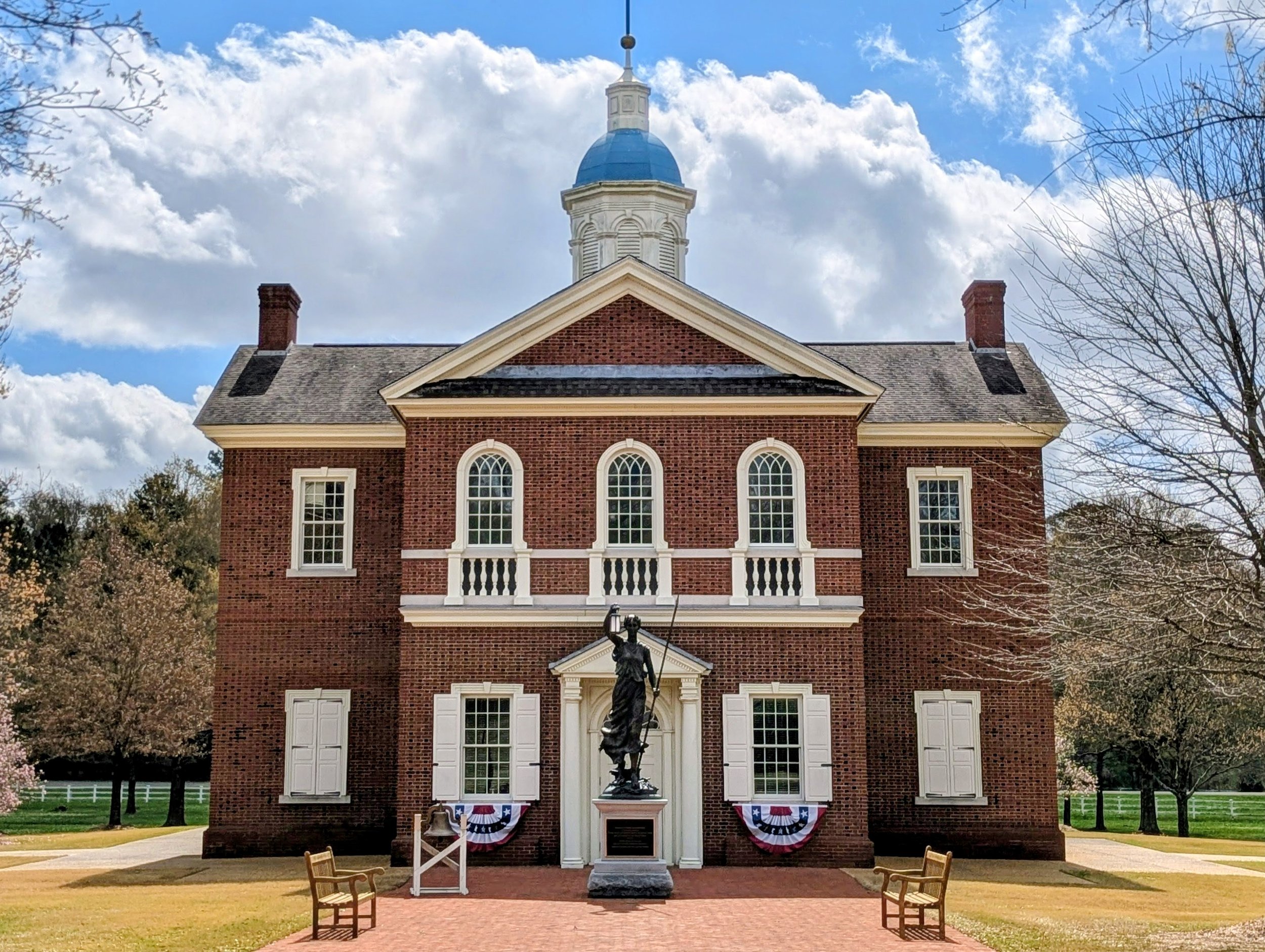 A historic red brick building with white accents and a blue domed cupola, featuring a statue of a woman holding a spear in front, surrounded by trees and a bright blue sky with clouds.
