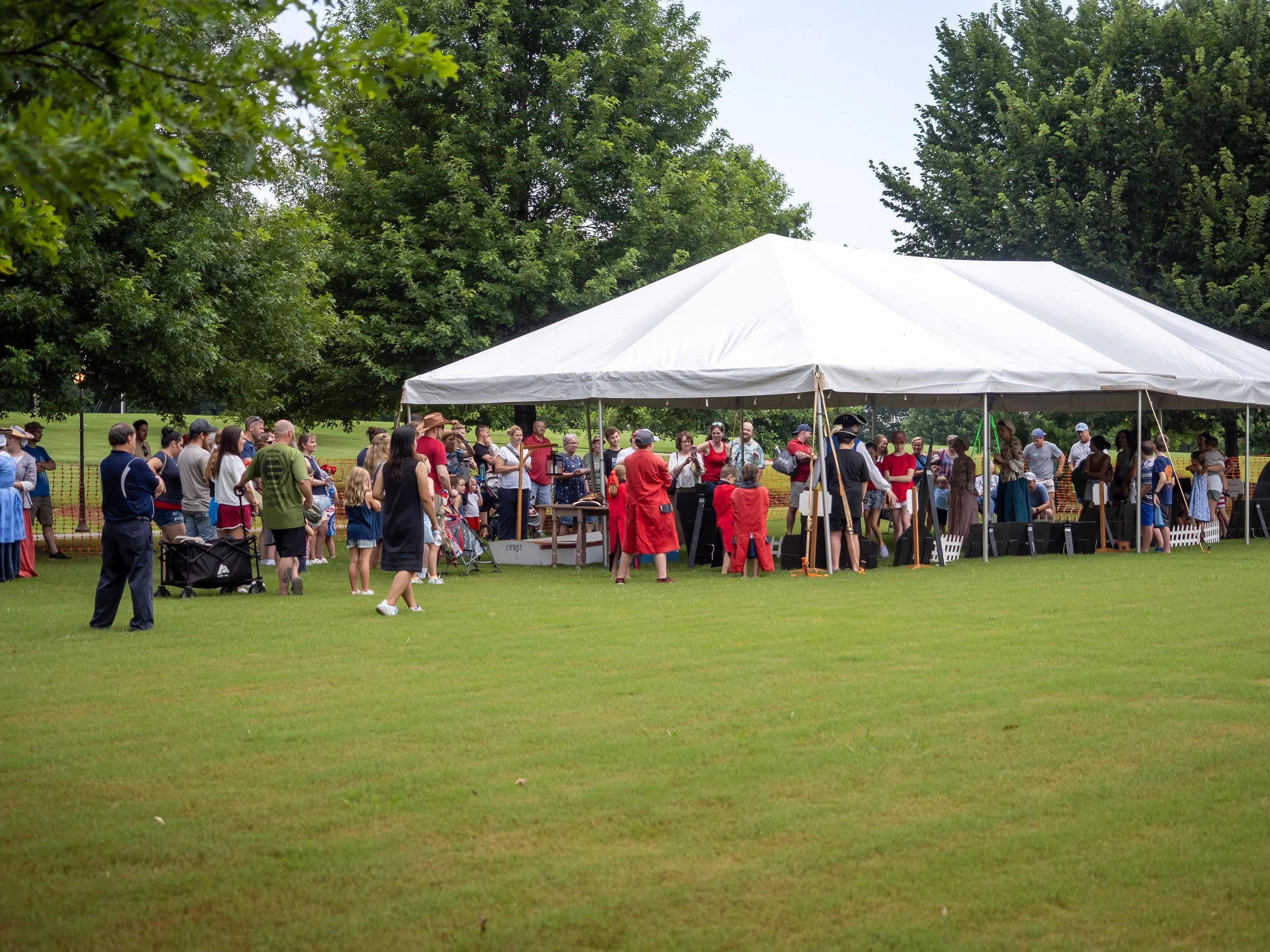 Outdoor gathering with many people under a large white event tent on a grassy field, surrounded by trees.