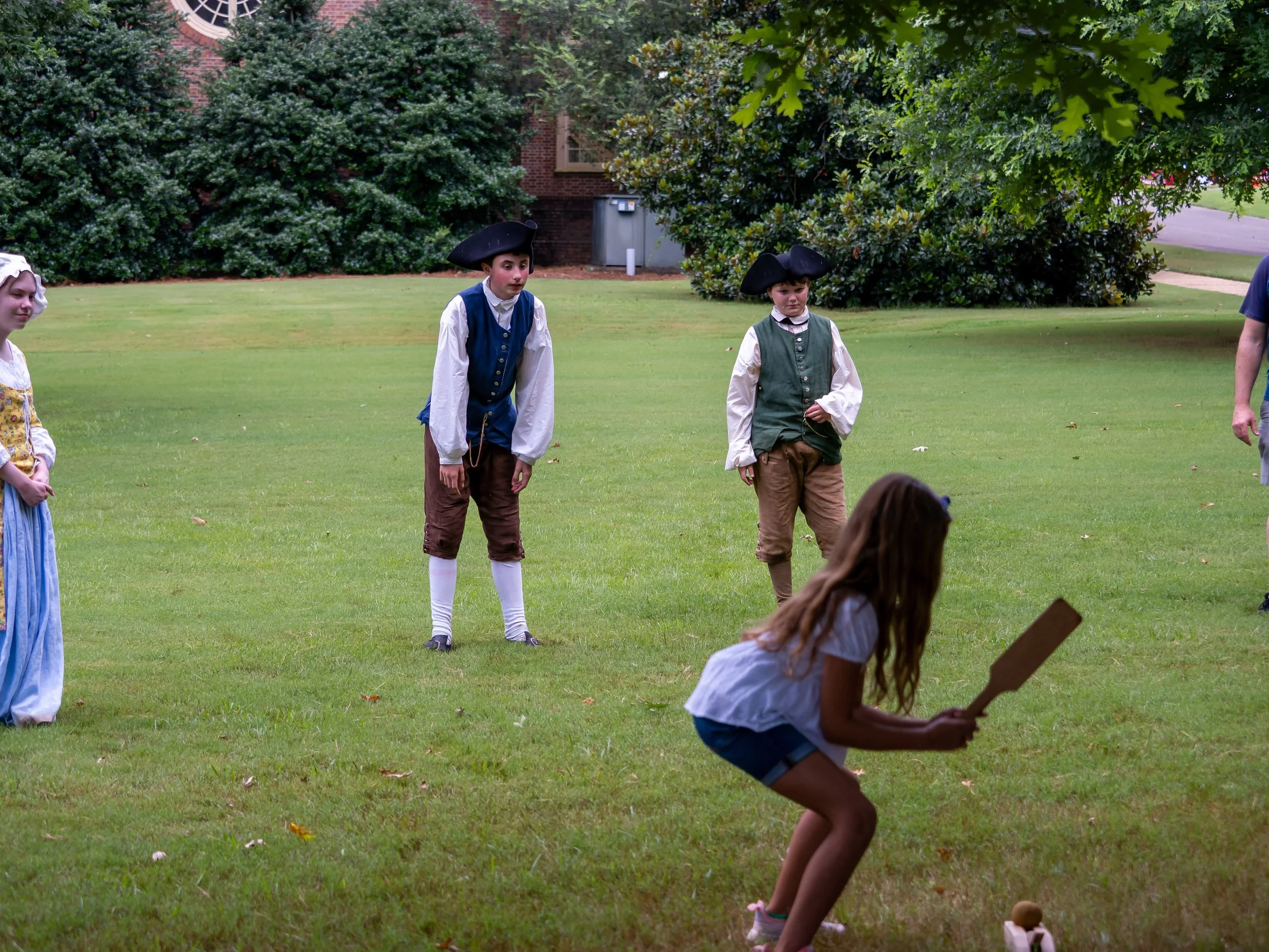 Children dressed in colonial costumes playing a game with a cricket bat on a grassy lawn.