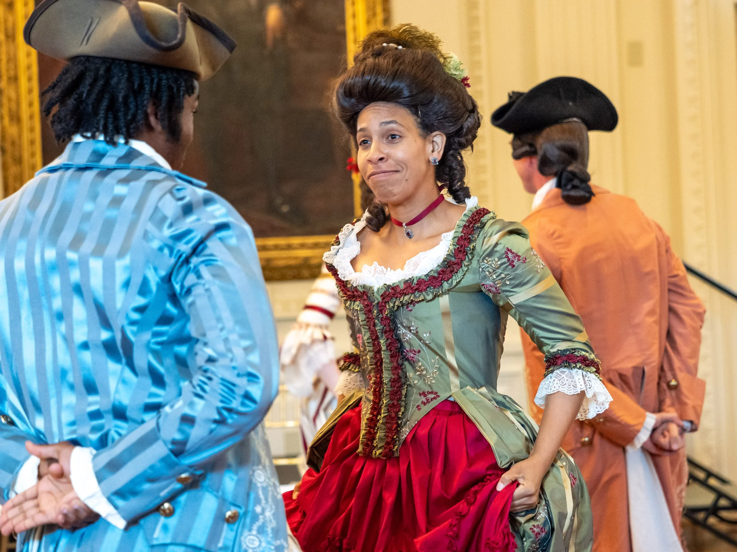 Women in 18th-century costumes, one in a green dress with red skirt talking to a woman in a blue striped dress, two men in 18th-century attire in the background inside a historic building.