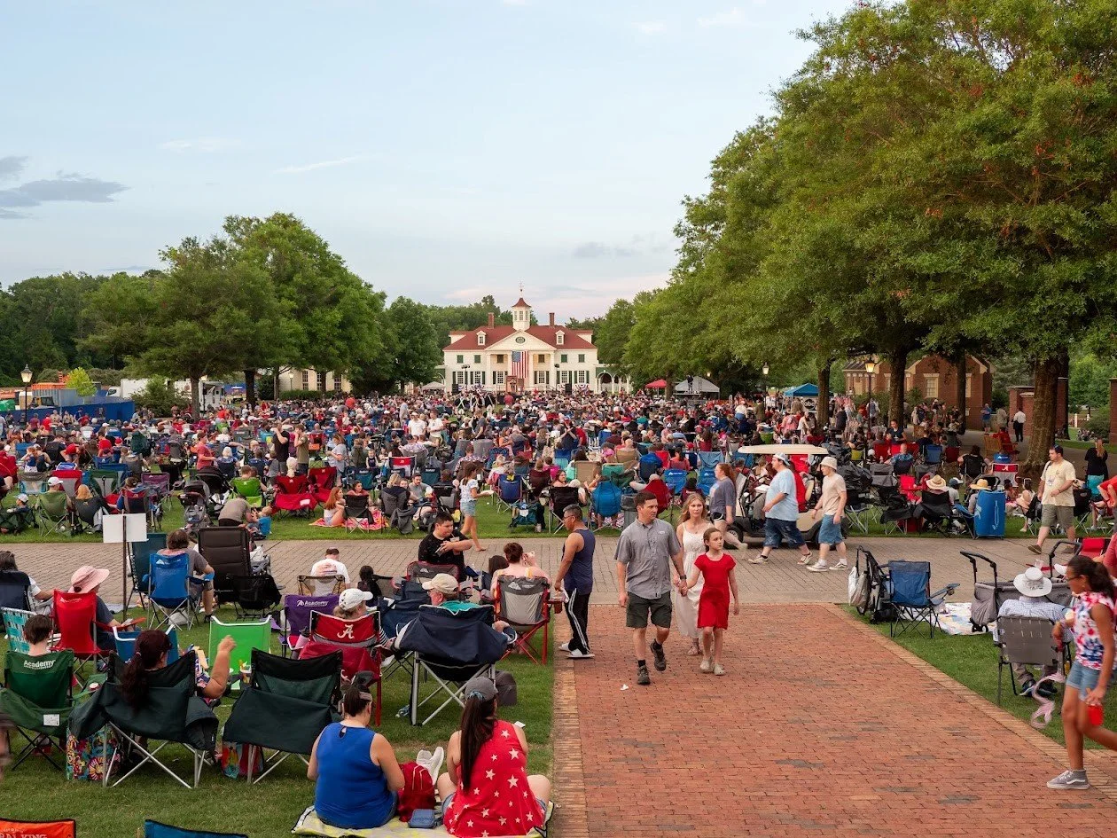 Crowd gathered on a lawn with chairs and blankets in front of a large white building with a clock tower, Red, white, and blue decorations, and American flags, likely for a community event or celebration.