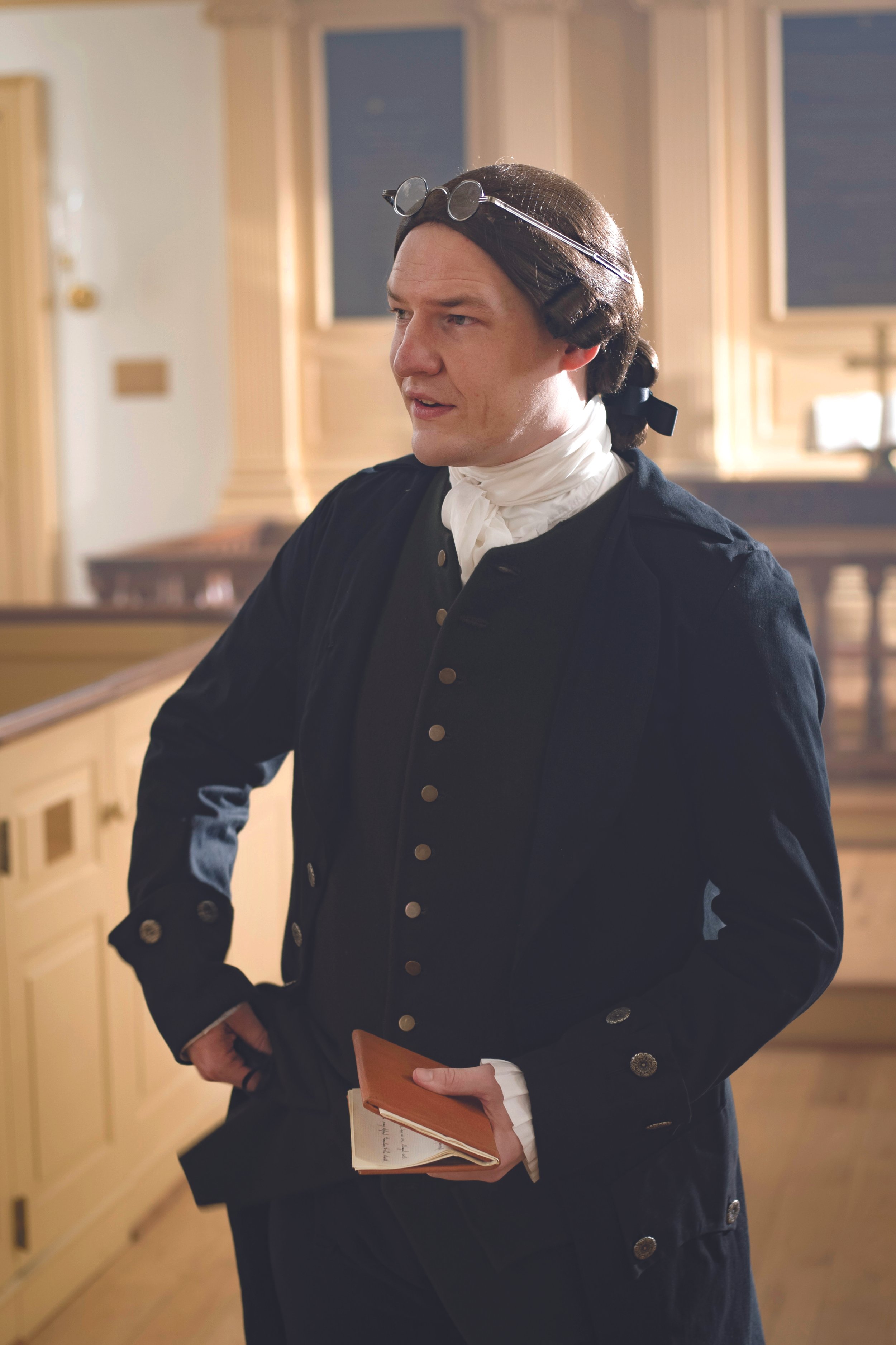 A man dressed in historical clothing, wearing a wig with a black ribbon, round glasses on his forehead, white neckcloth, black coat with buttons, holding a small leather-bound book in his left hand, standing in a wood-paneled room with blue windows.