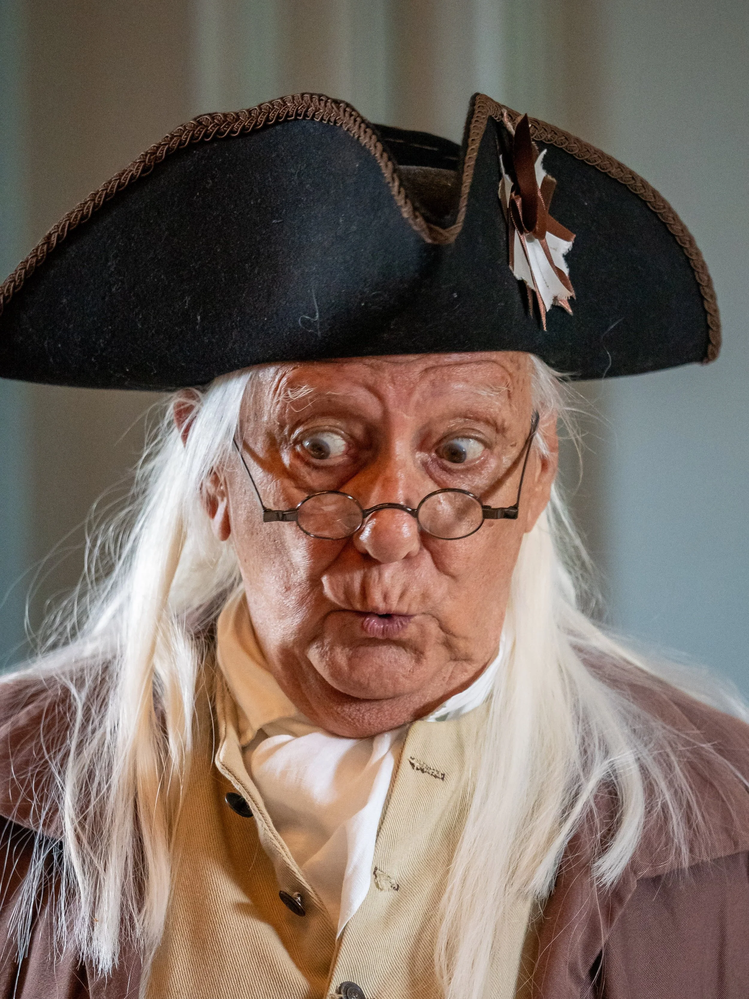 A man dressed as a historical figure, wearing a colonial-style coat and a large black tricorn hat with brown trimmings, round spectacles, and long white hair.