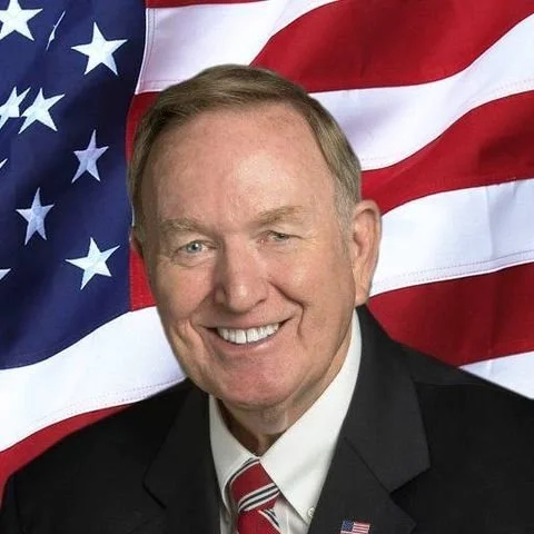 Sen. Tom Butler in a suit and tie stands in front of an American flag.