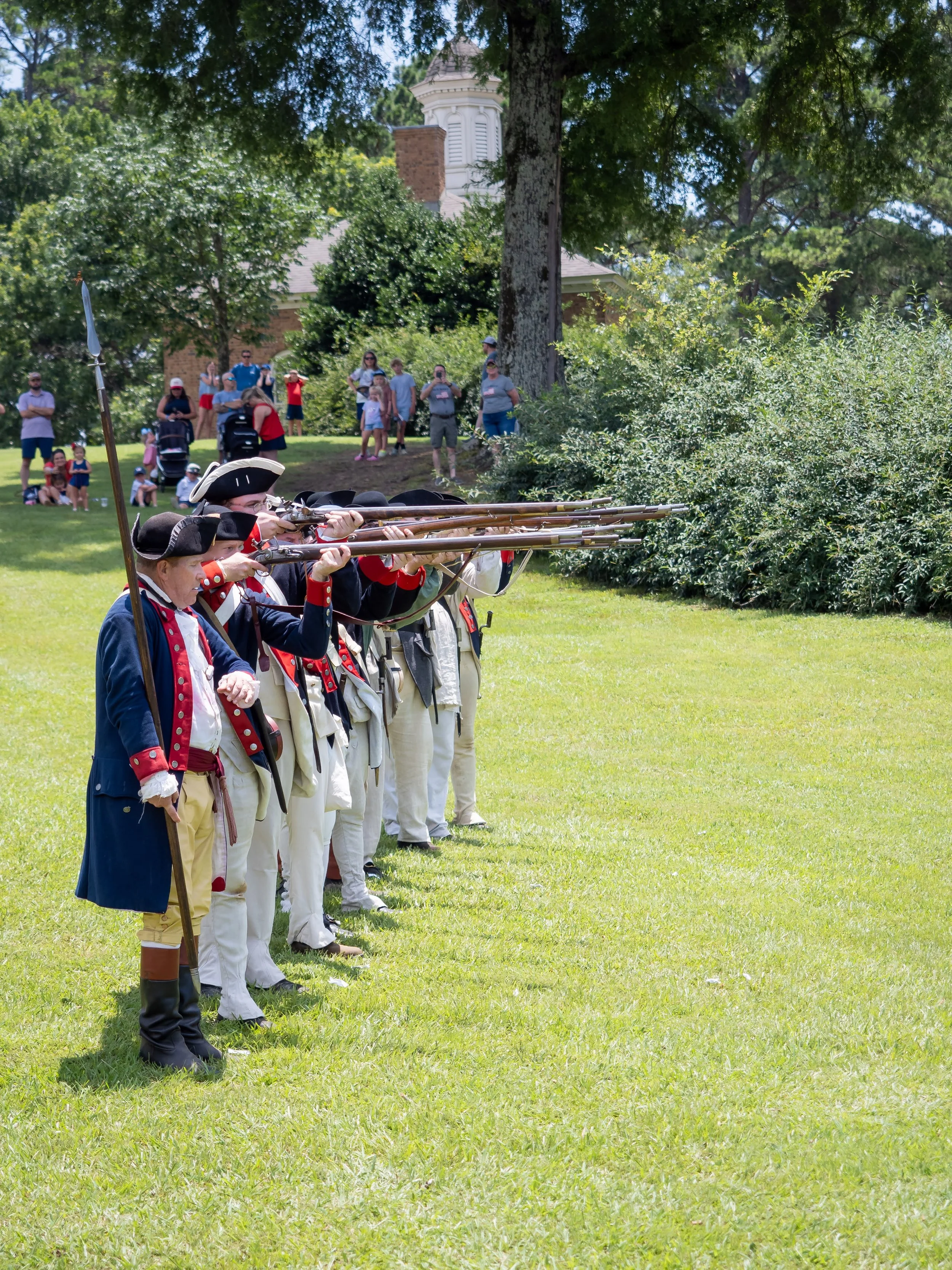 Reenactors dressed in 18th-century military uniforms aiming muskets during a historical reenactment in a park, with spectators watching in the background.