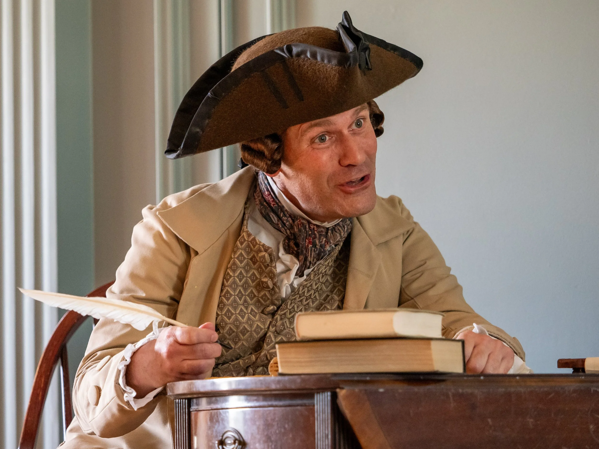 A man in historical costume sitting at a desk, holding a quill, wearing a large tricorn hat and a beige coat, appearing to speak or explain something.