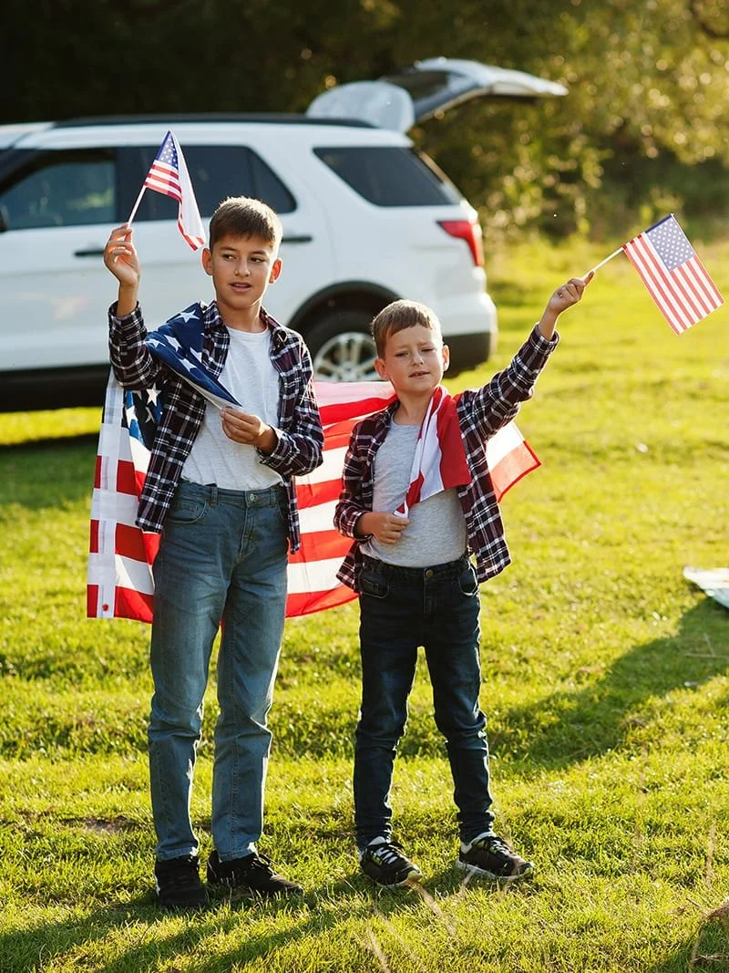 Two boys holding small American flags outside near a car with the trunk open. They are draped in American flags and seem to be celebrating, possibly at a patriotic event, on a bright, sunny day.