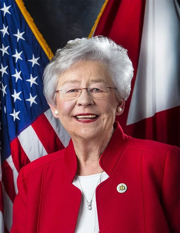 Gov. Kay Ivey wearing a red blazer and a white top, standing in front of American and another flag, smiling at the camera.