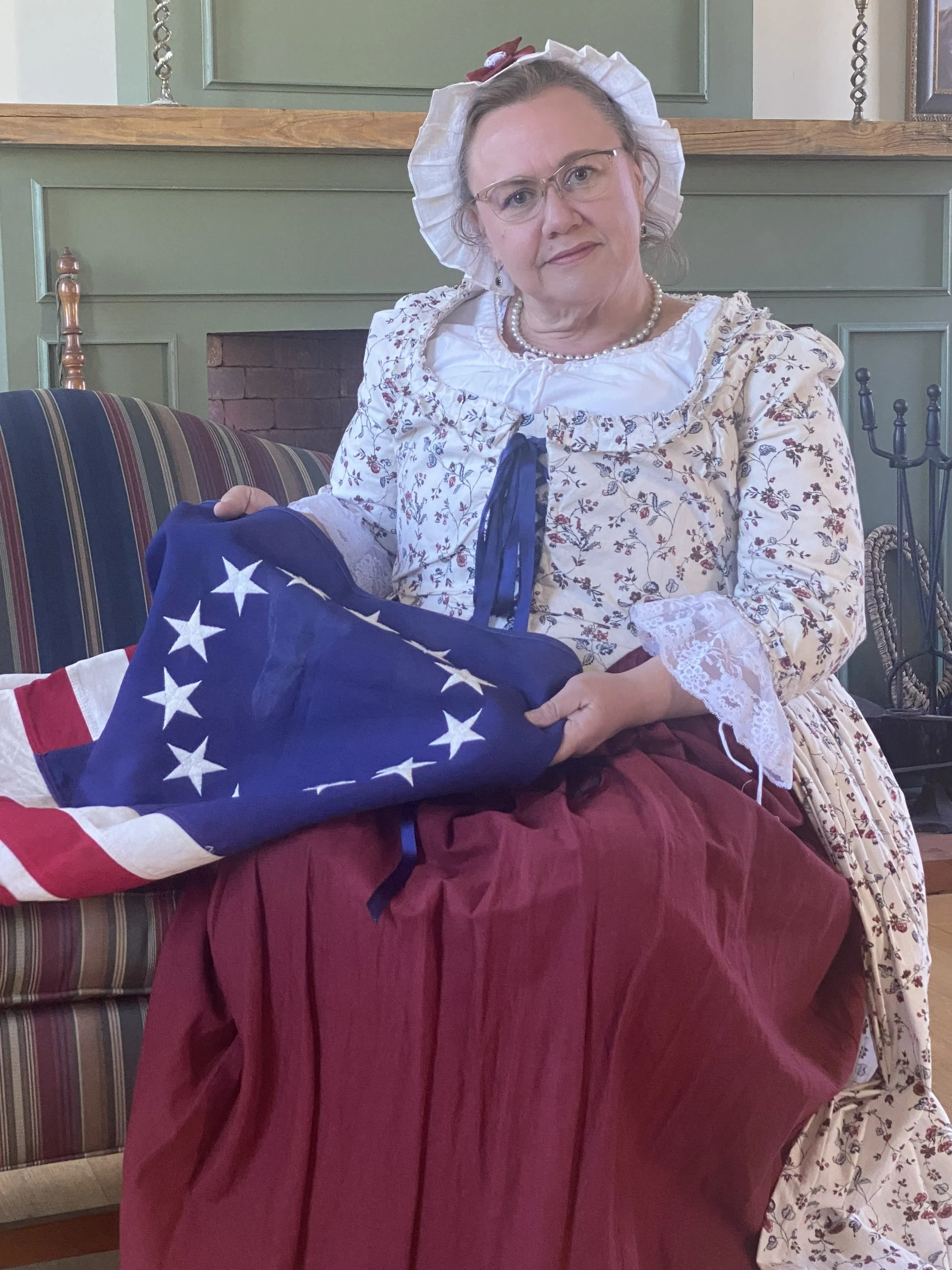 A woman dressed in historical clothing, including a bonnet and lace-trimmed dress, sitting on a couch and holding a folded American flag.