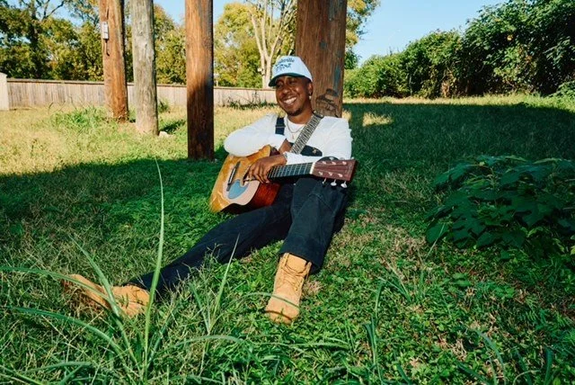 Michael Warren sitting on grass under trees, holding an acoustic guitar, smiling, wearing a white shirt, black pants, tan boots, and a cap, in a backyard setting.