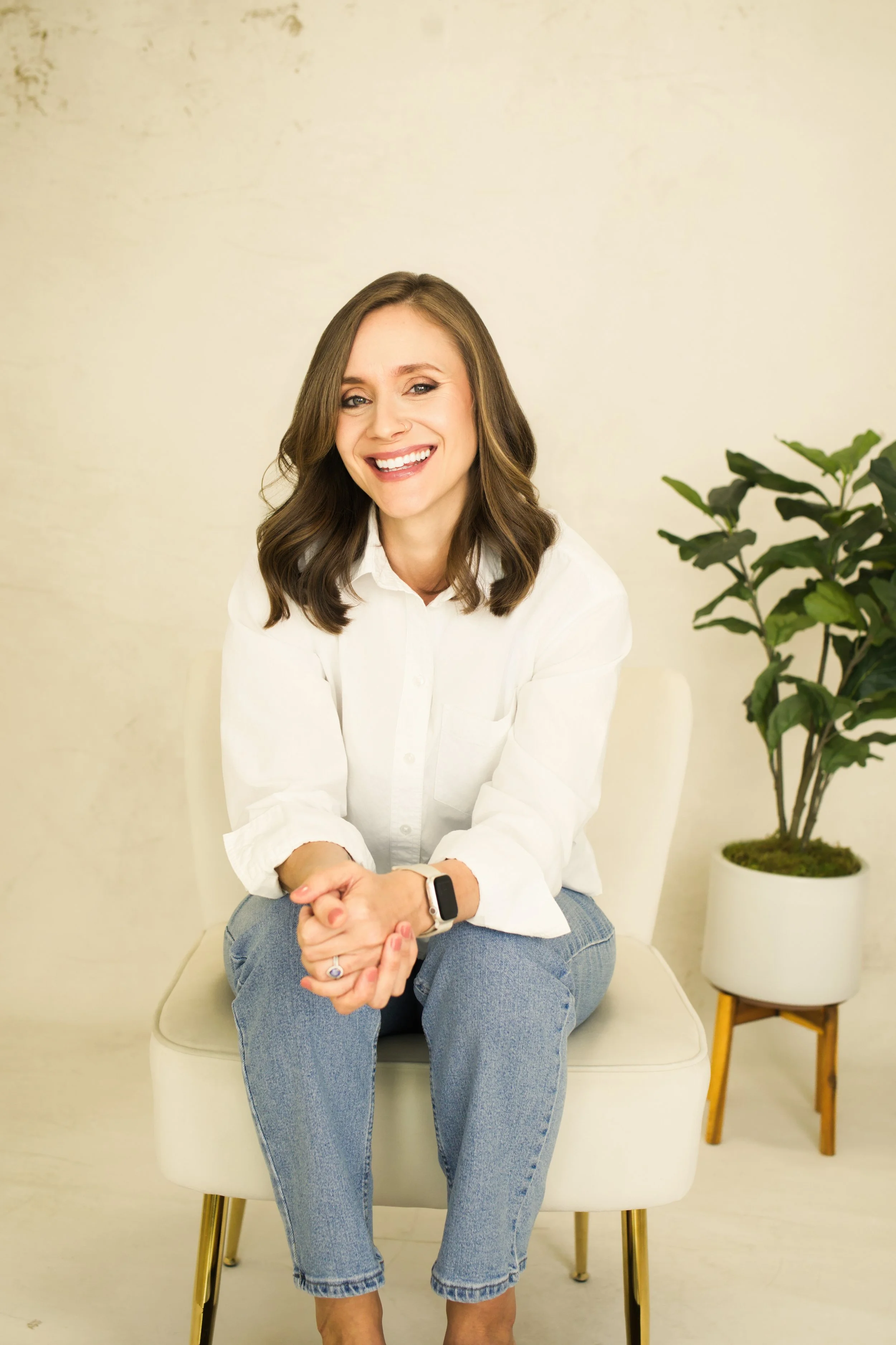 Tara Gogolinski, couples therapist in Raleigh, NC, sitting on a white chair and smiling at the camera in a white shirt and blue jeans, with a potted plant behind her.