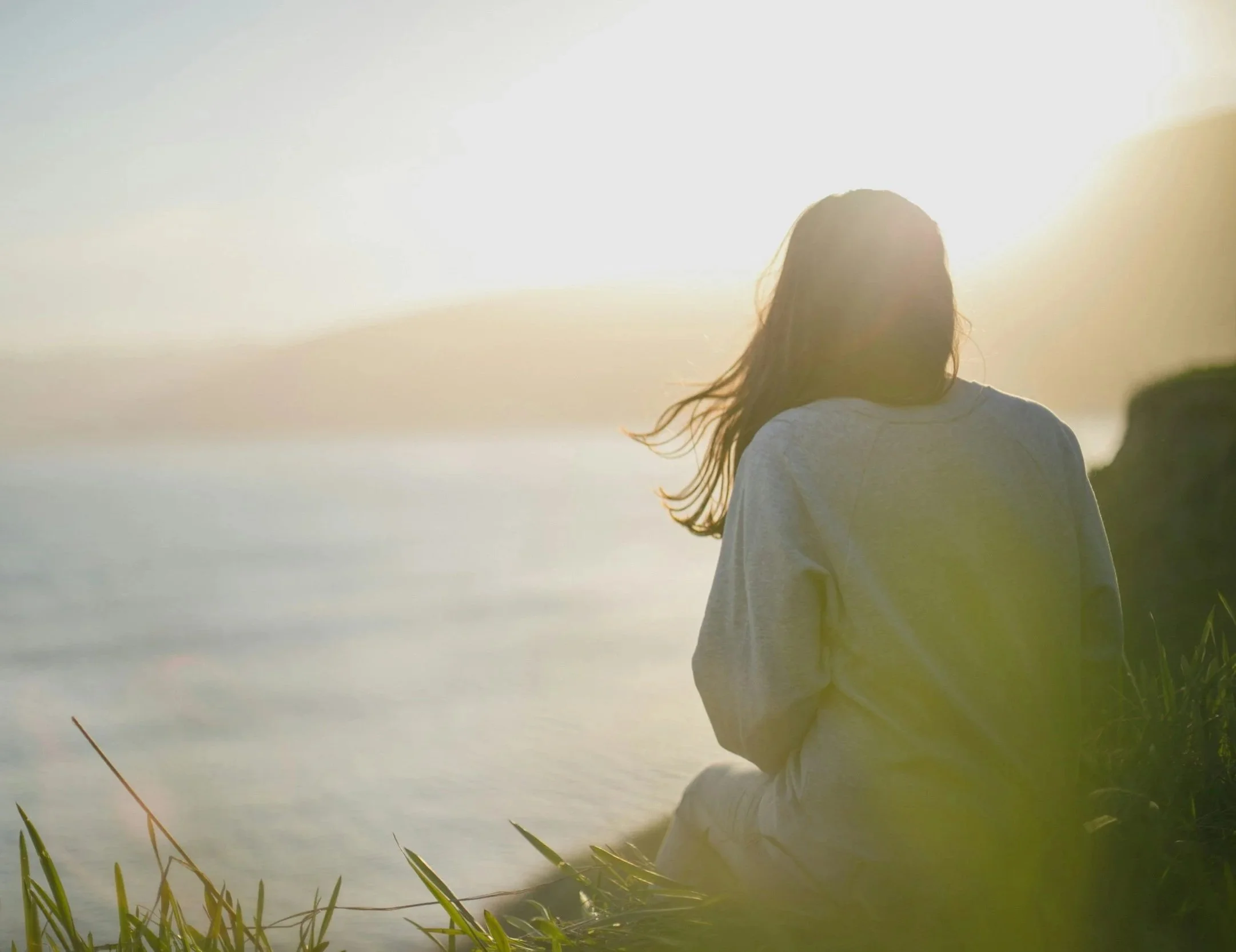 A person sitting outdoors near a body of water during sunset, facing away from the camera with long hair blowing in the wind.