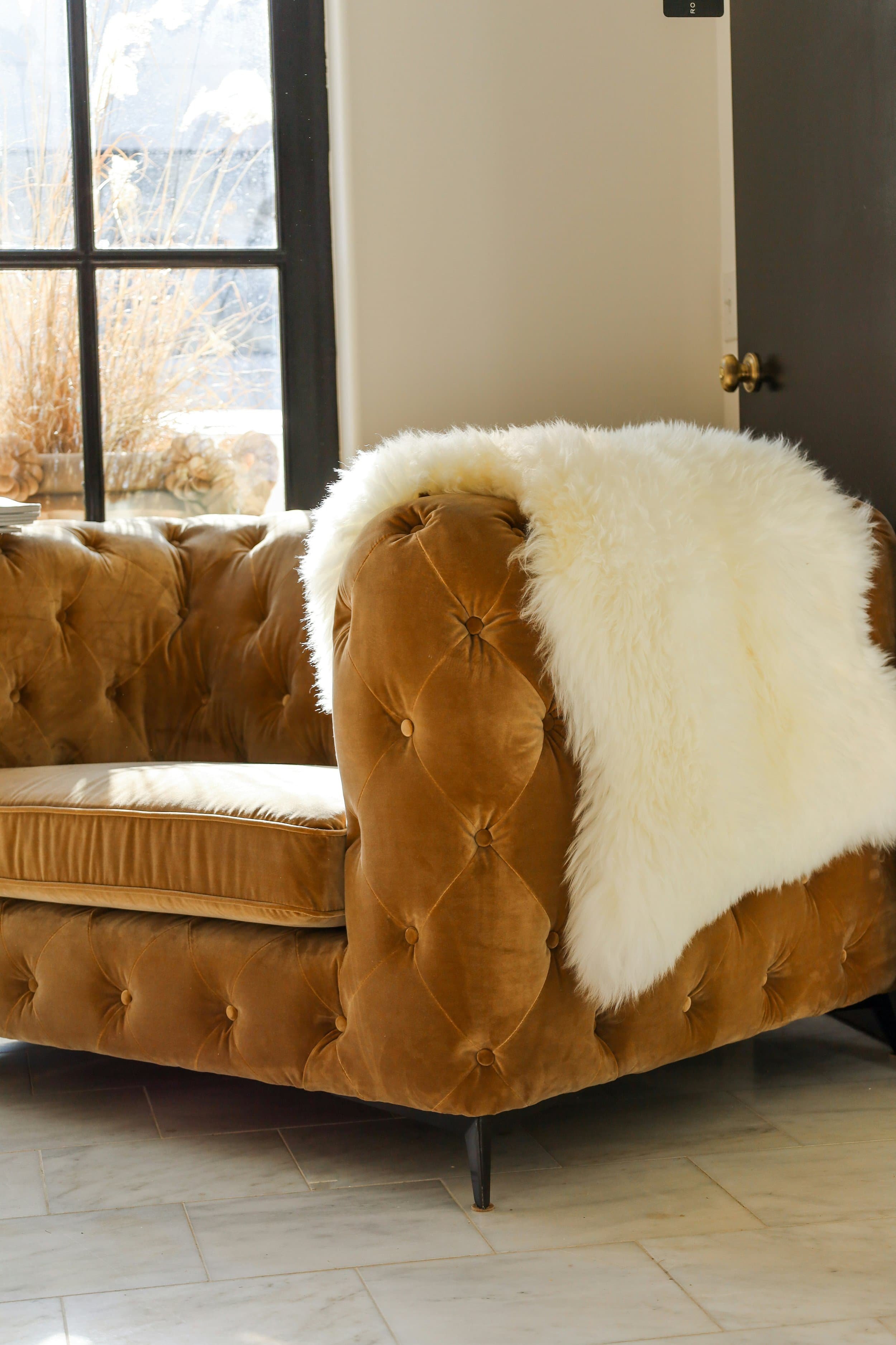 A brown tufted velvet sofa with a white faux fur throw blanket near a sunlit window, symbolizing the calm, welcoming space offered through therapy services in Raleigh, NC.