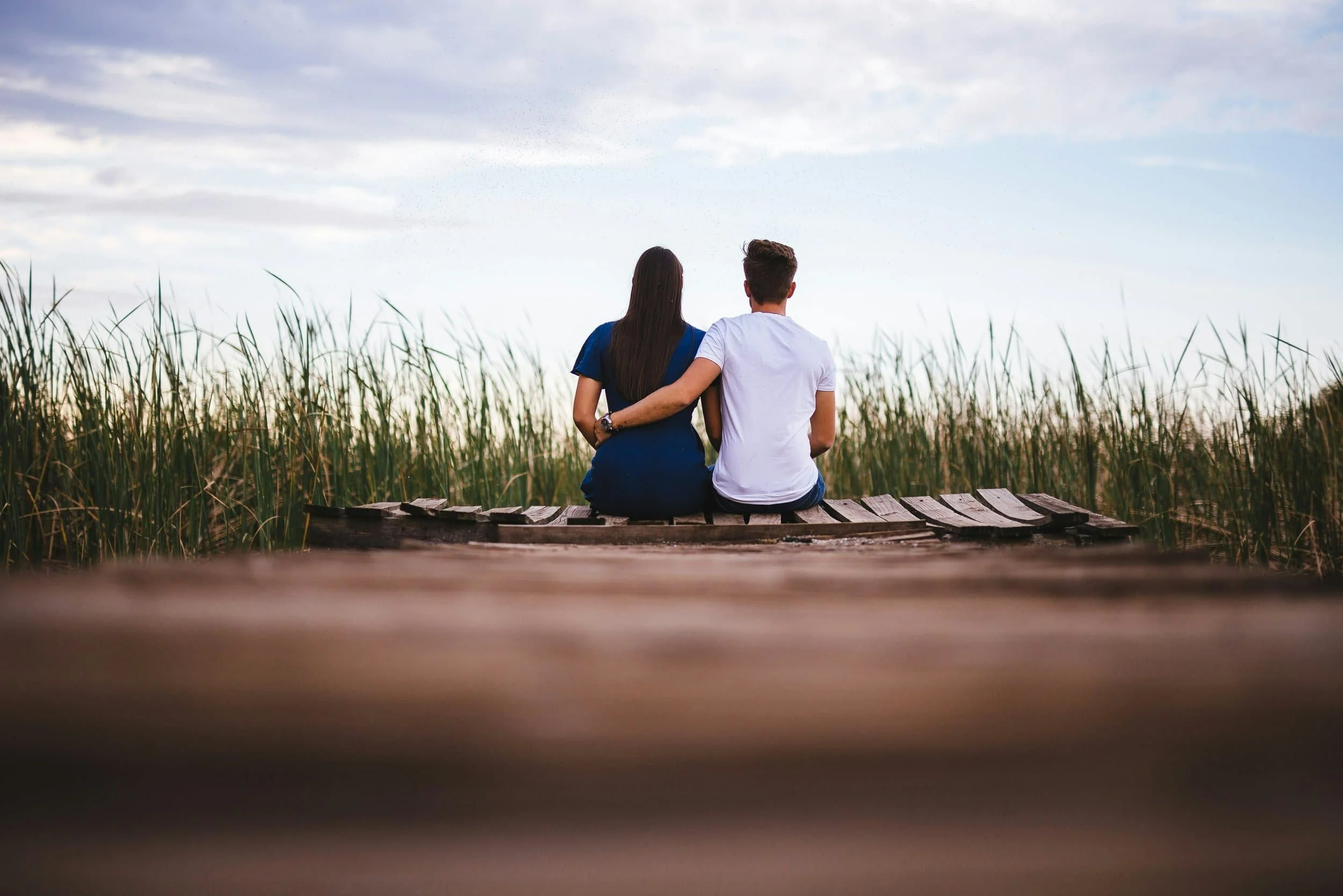 A couple sitting on a wooden dock at the edge of a field with tall grass, watching a cloudy sky at sunset or sunrise.