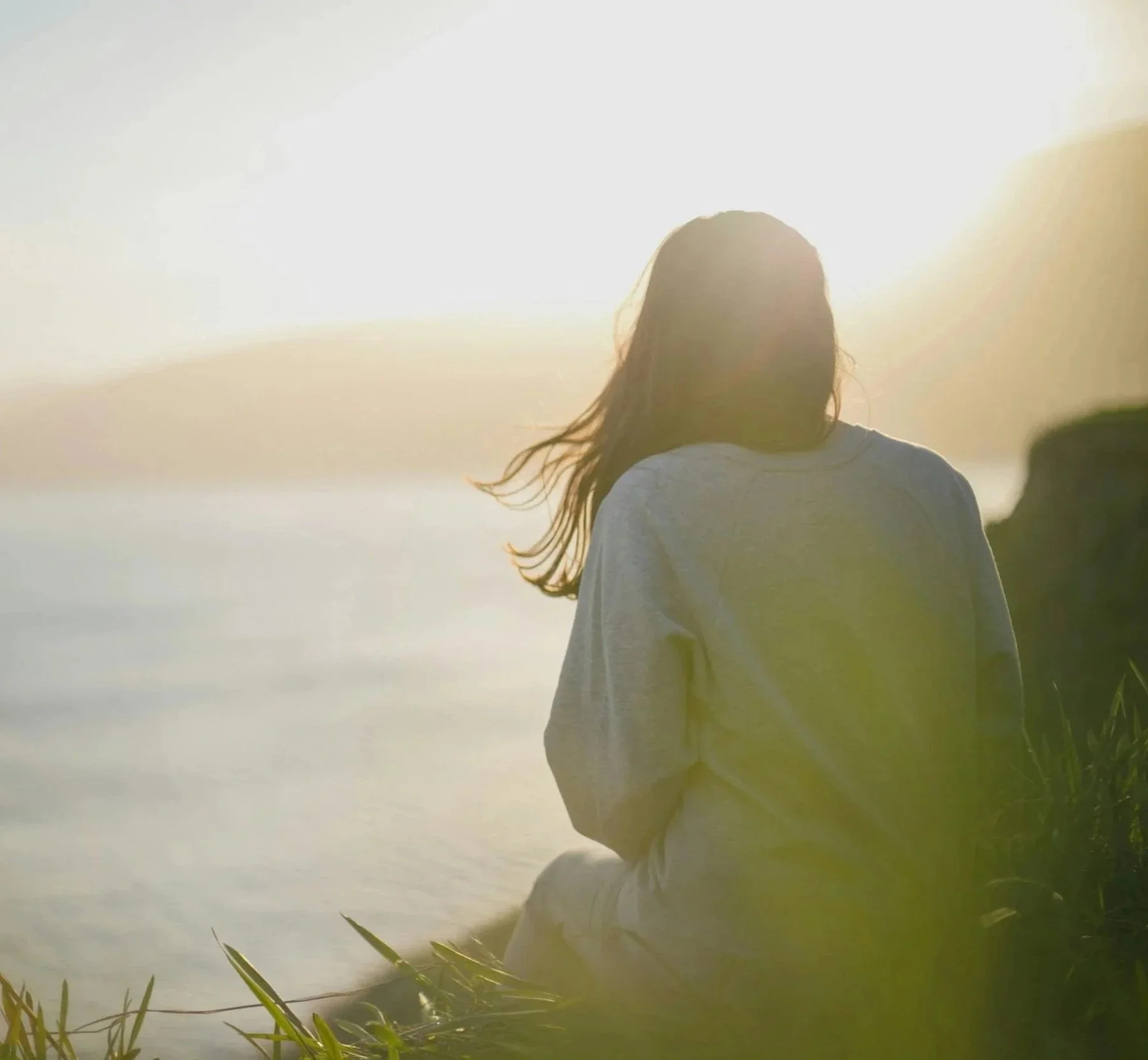 A woman sitting on the grass by the water at sunset, facing away from the camera, symbolizing the personal reflection and healing offered through individual therapy in Raleigh, NC.