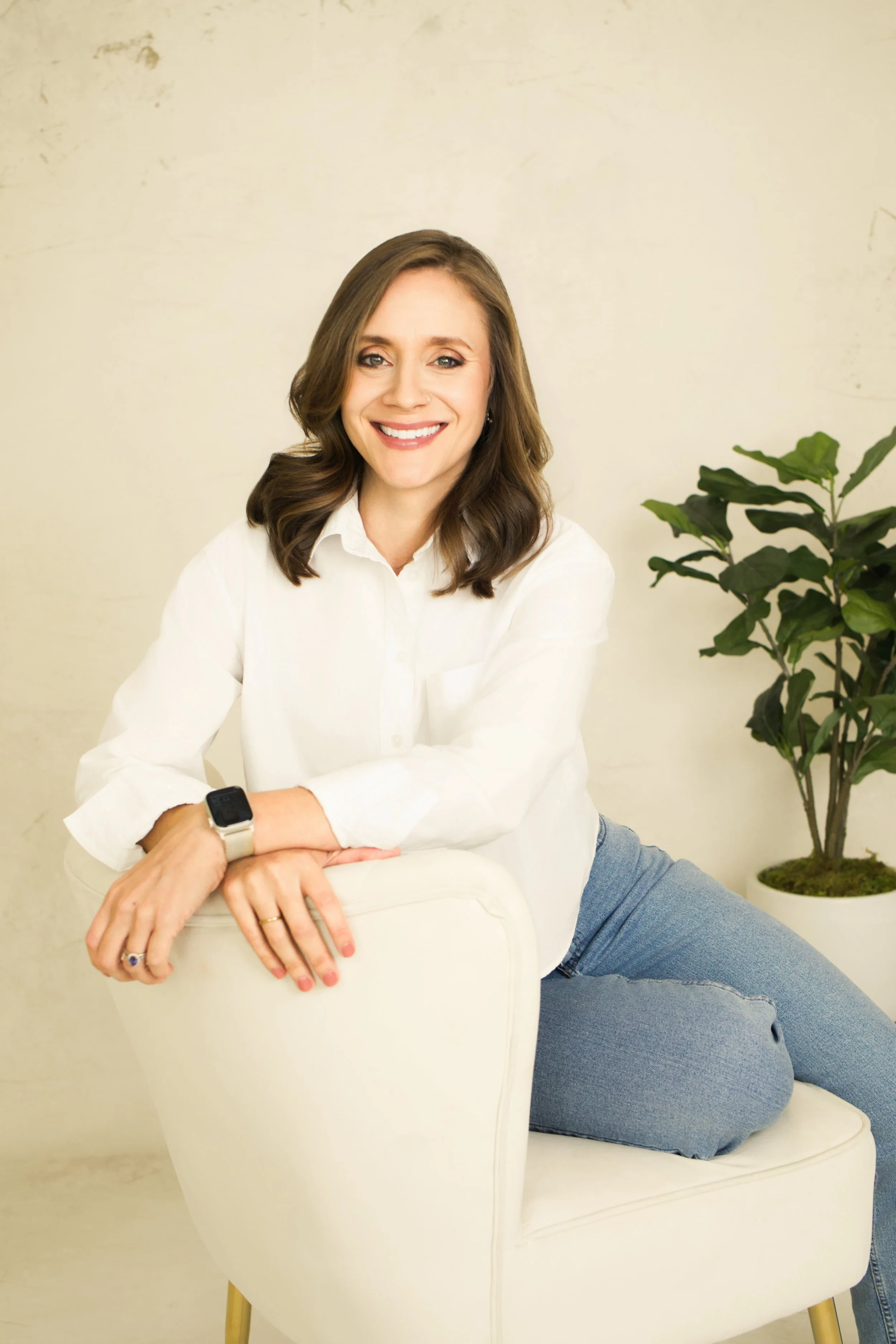 Tara Gogolinski, couples therapist in Raleigh, NC, smiling while seated on a white armchair in a white shirt and jeans, wearing a smartwatch and ring, with a green potted plant behind her.
