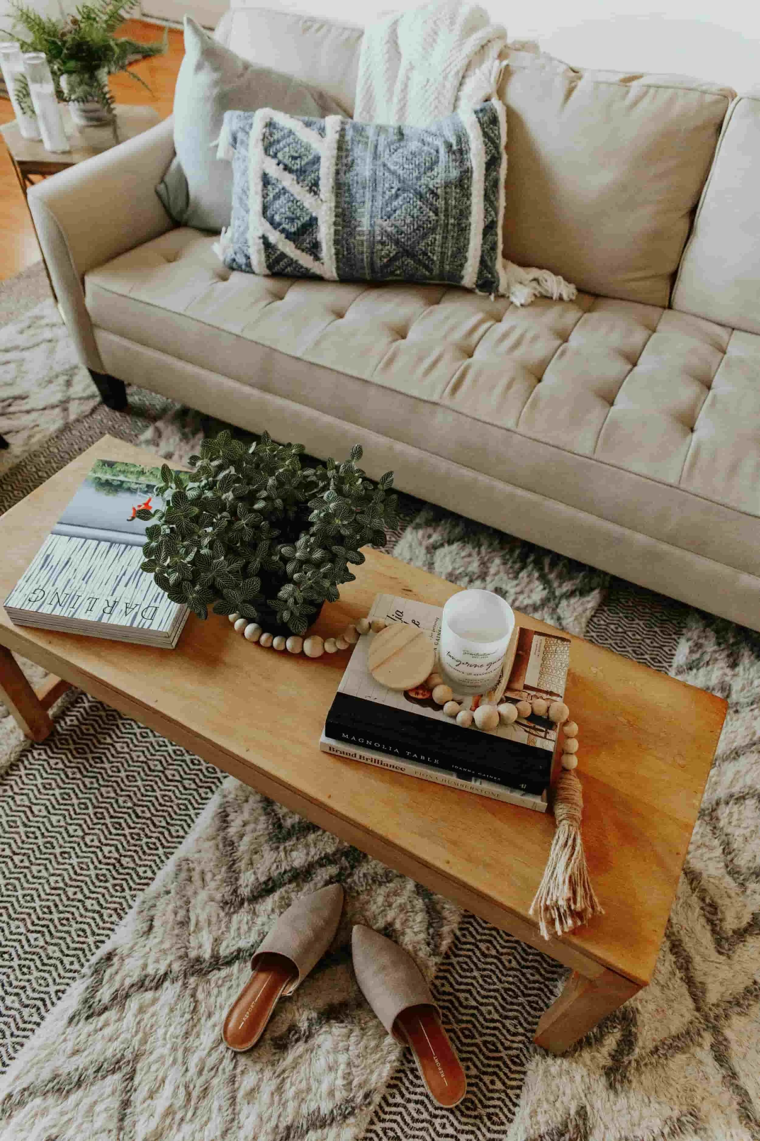 Living room with a beige tufted sofa, decorative pillows, a wooden coffee table with books, a candle, a small wooden box, a beaded garland, and a plant, with patterned rugs and shoes on the floor.
