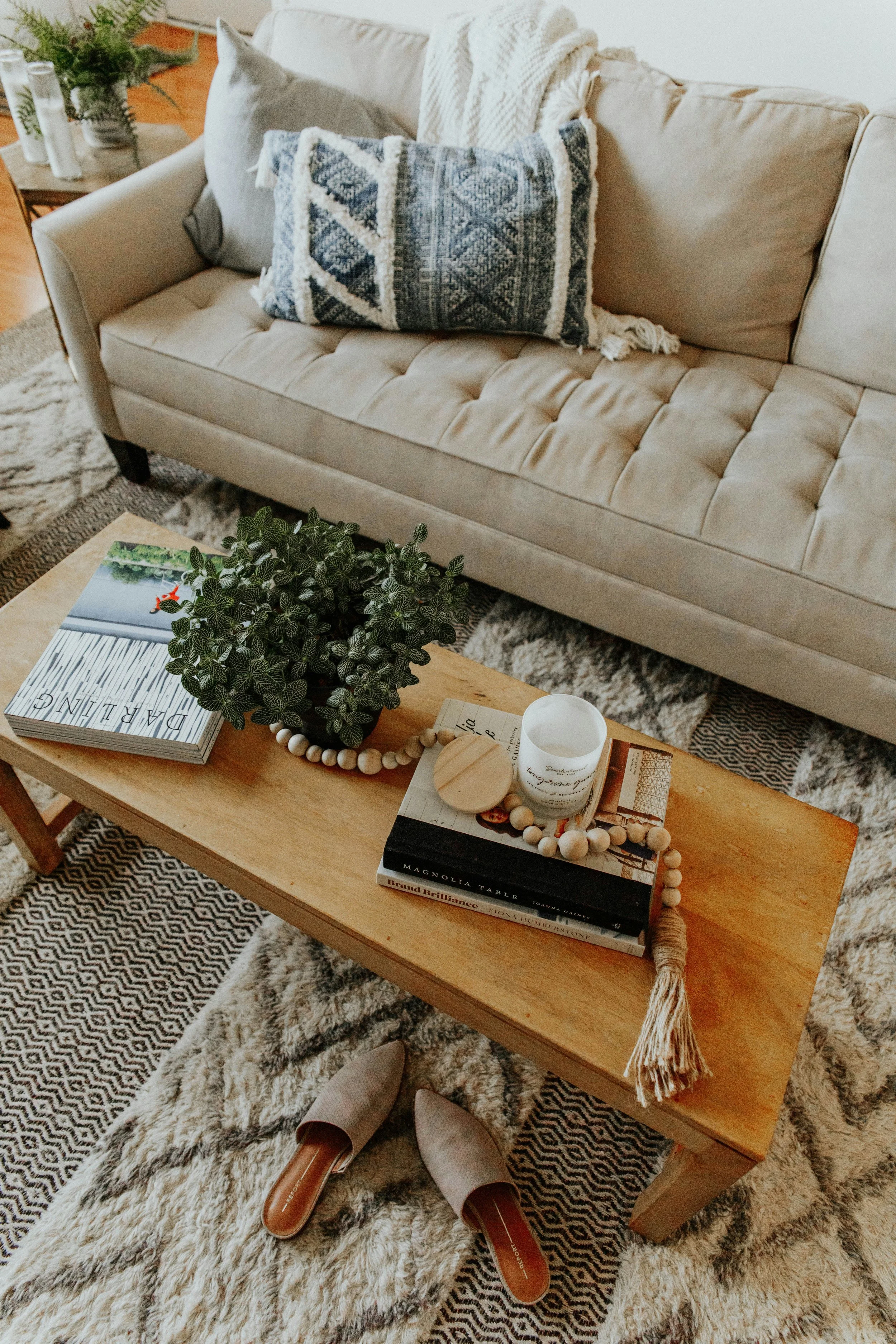 Living room with a beige tufted sofa, decorative pillows, a wooden coffee table with books, a candle, a small wooden box, a beaded garland, and a plant, with patterned rugs and shoes on the floor.