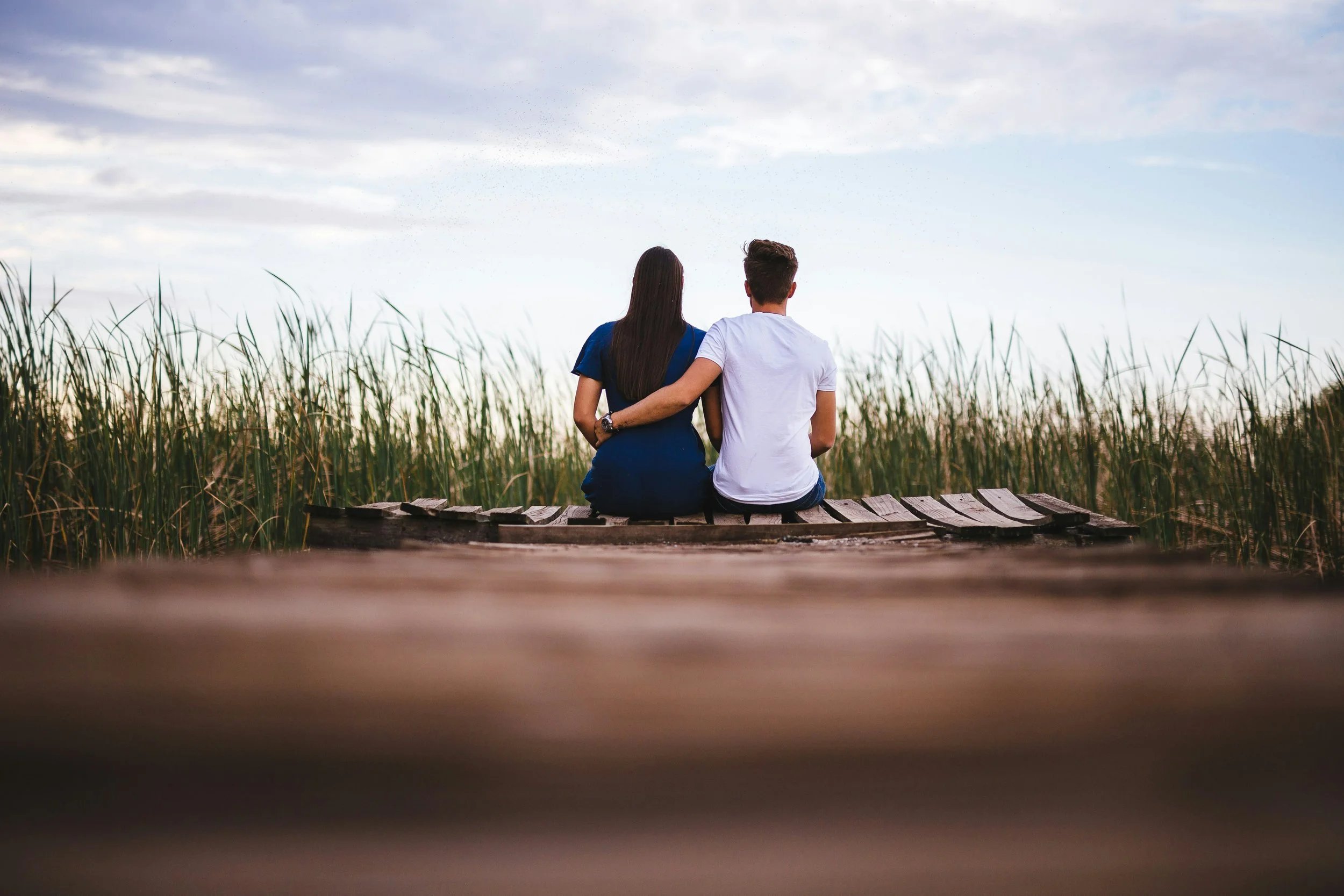 A couple sitting on a wooden dock at the edge of a body of water, facing away from the camera, with tall grass or reeds surrounding them and a cloudy sky overhead.
