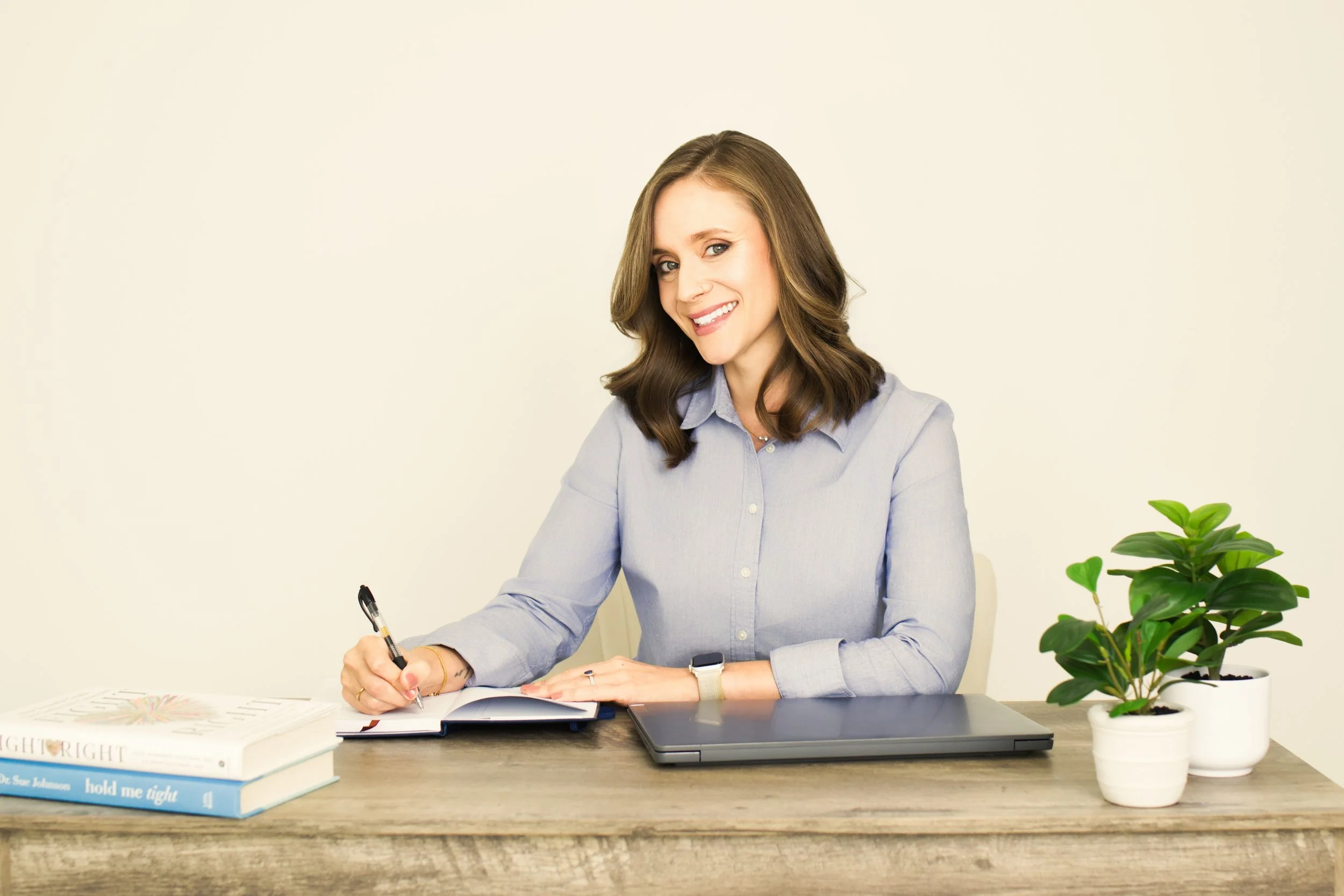 Tara Gogolinski, couples therapist in Raleigh, NC, smiling while taking notes at a wooden desk with plants, books, a closed laptop, and a notebook, reflecting her warm, insightful approach to therapy.