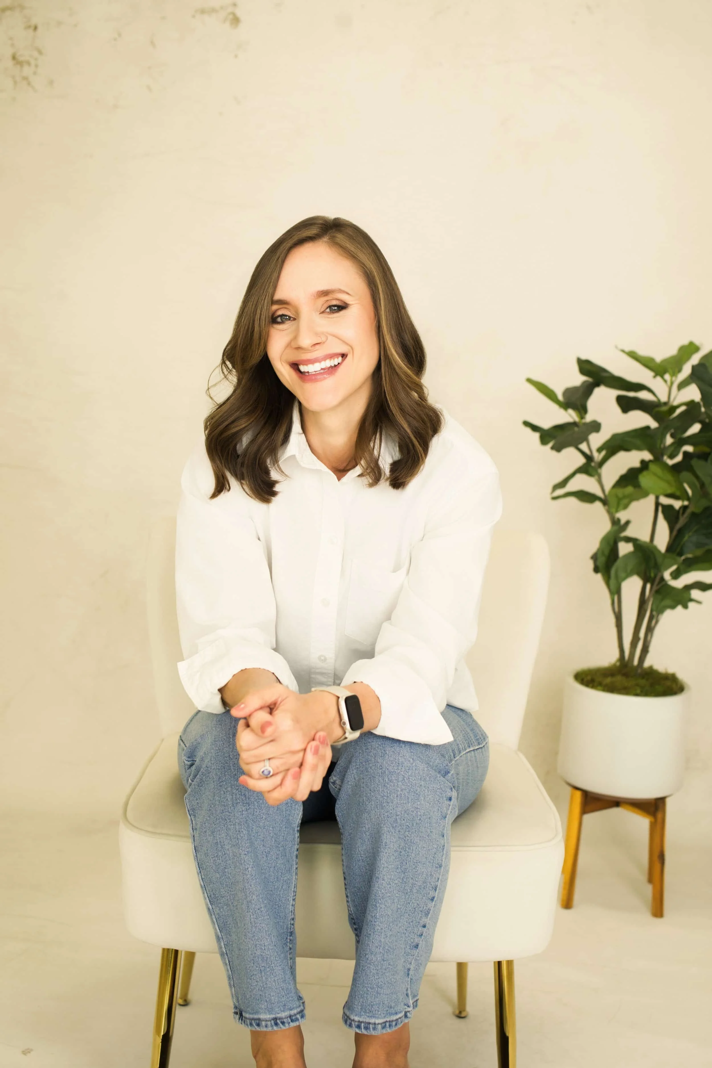Tara Gogolinski, couples therapist in Raleigh, NC, sitting on a white chair and smiling at the camera in a white shirt and blue jeans, with a potted plant behind her.