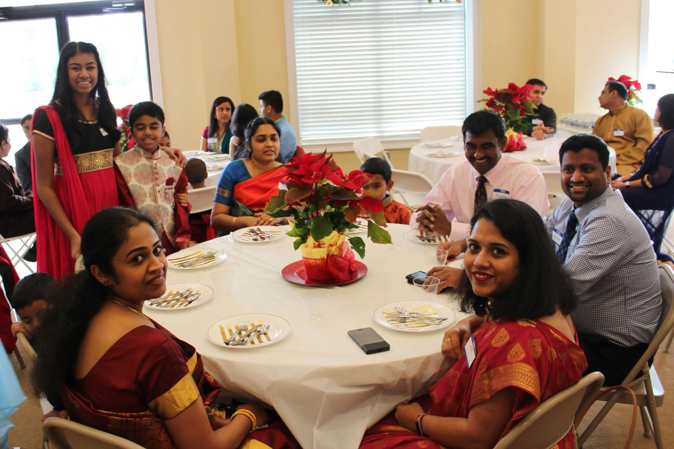 Group of people celebrating a gathering at a decorated table with a poinsettia centerpiece, some dressed in traditional South Asian attire, smiling and posing for the photo inside a well-lit room.