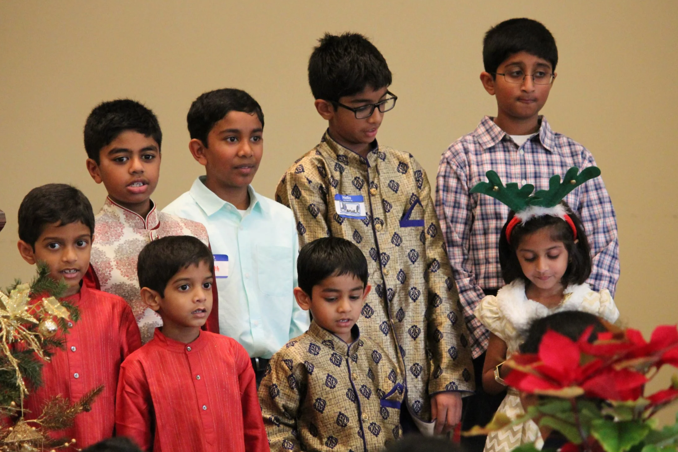 Group of children dressed in festive clothing at holiday event, one girl wearing reindeer antlers headband, some children with name tags.