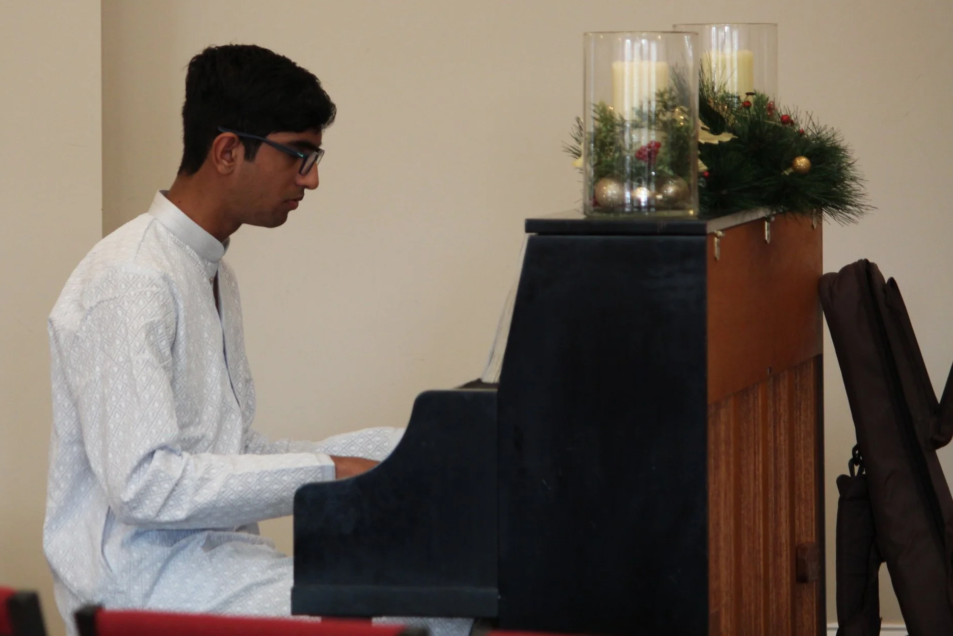 Man dressed in white traditional attire playing a black upright piano, decorated with Christmas ornaments and candles on top.