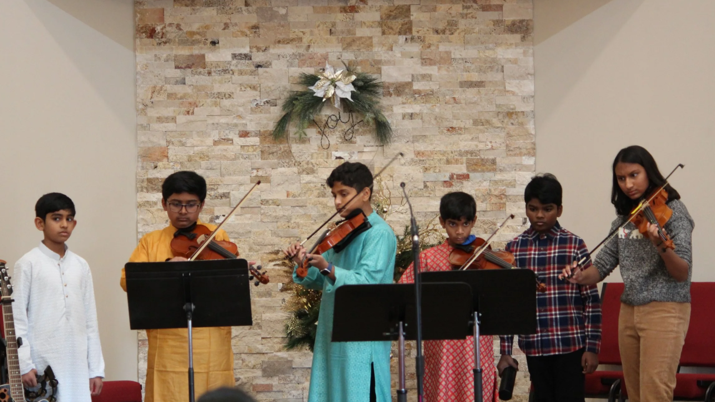 Group of six children playing violins and one girl standing in front of a festive Christmas decoration on a brick wall.
