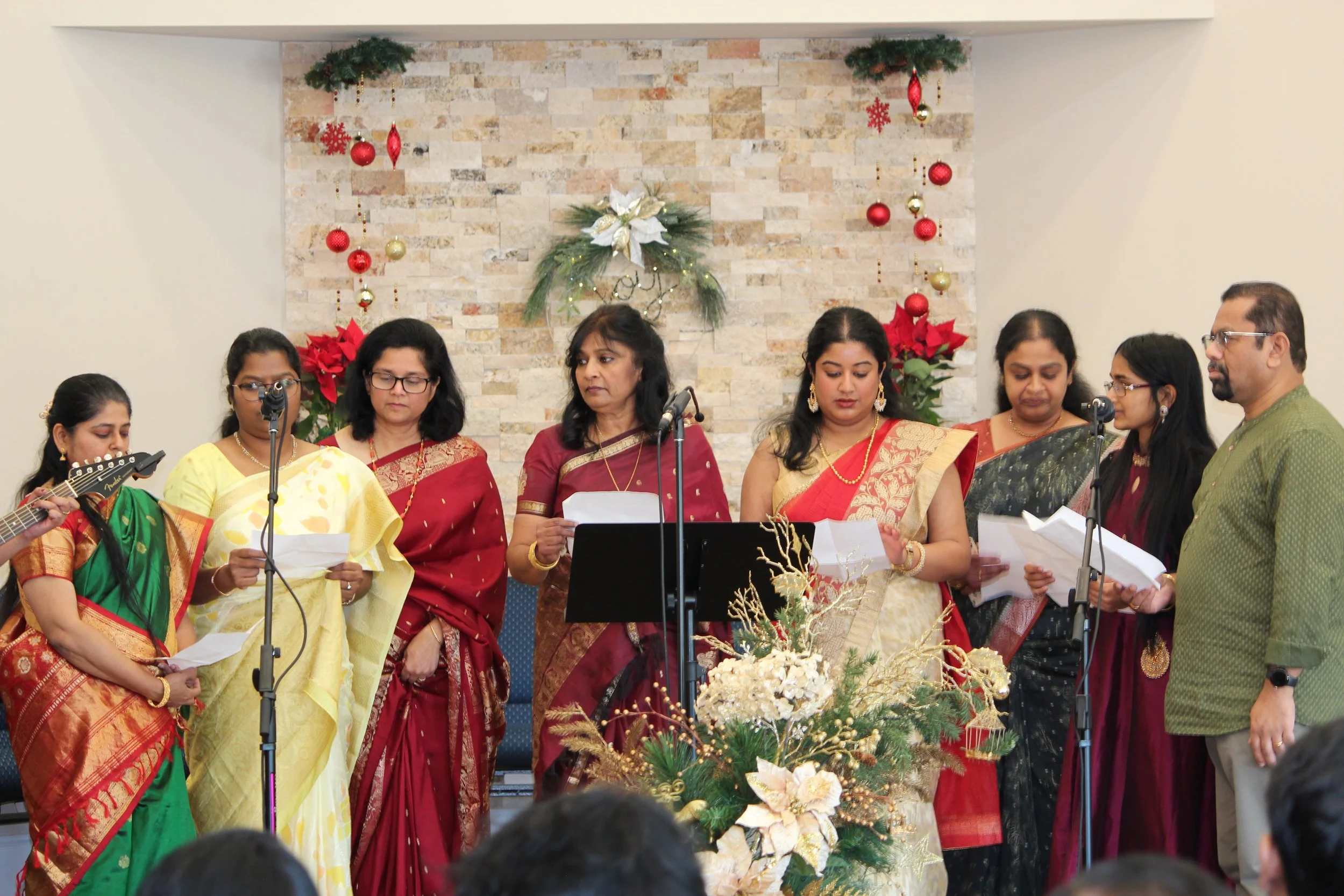 Group of people dressed in traditional Indian attire standing on a stage decorated with Christmas ornaments, flowers, and greenery, holding papers and singing or speaking.