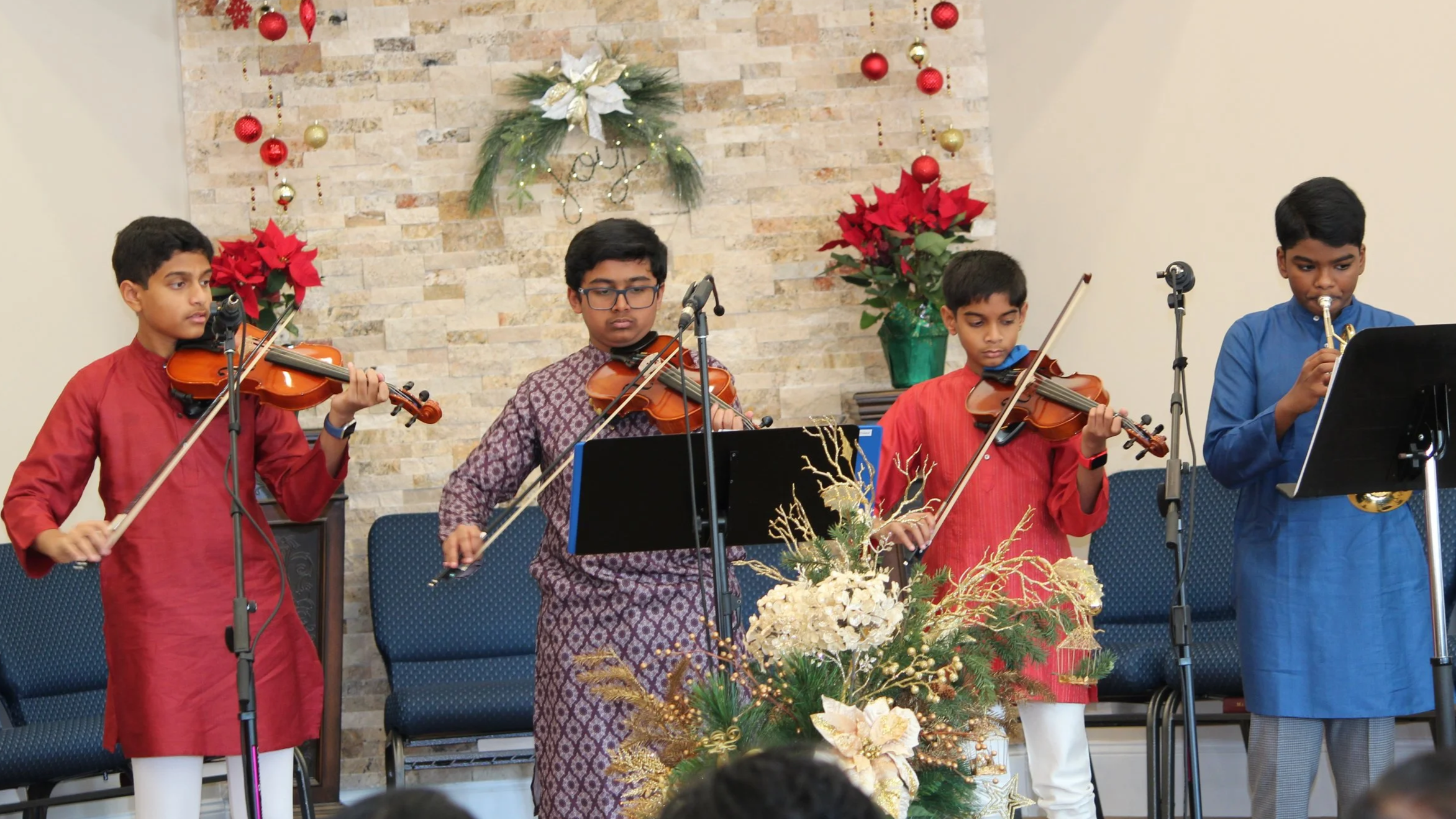 Four boys playing violins and a trumpet during a Christmas performance, decorated with poinsettias, poinsettia and Christmas ornaments, on a stage with a brick wall background.
