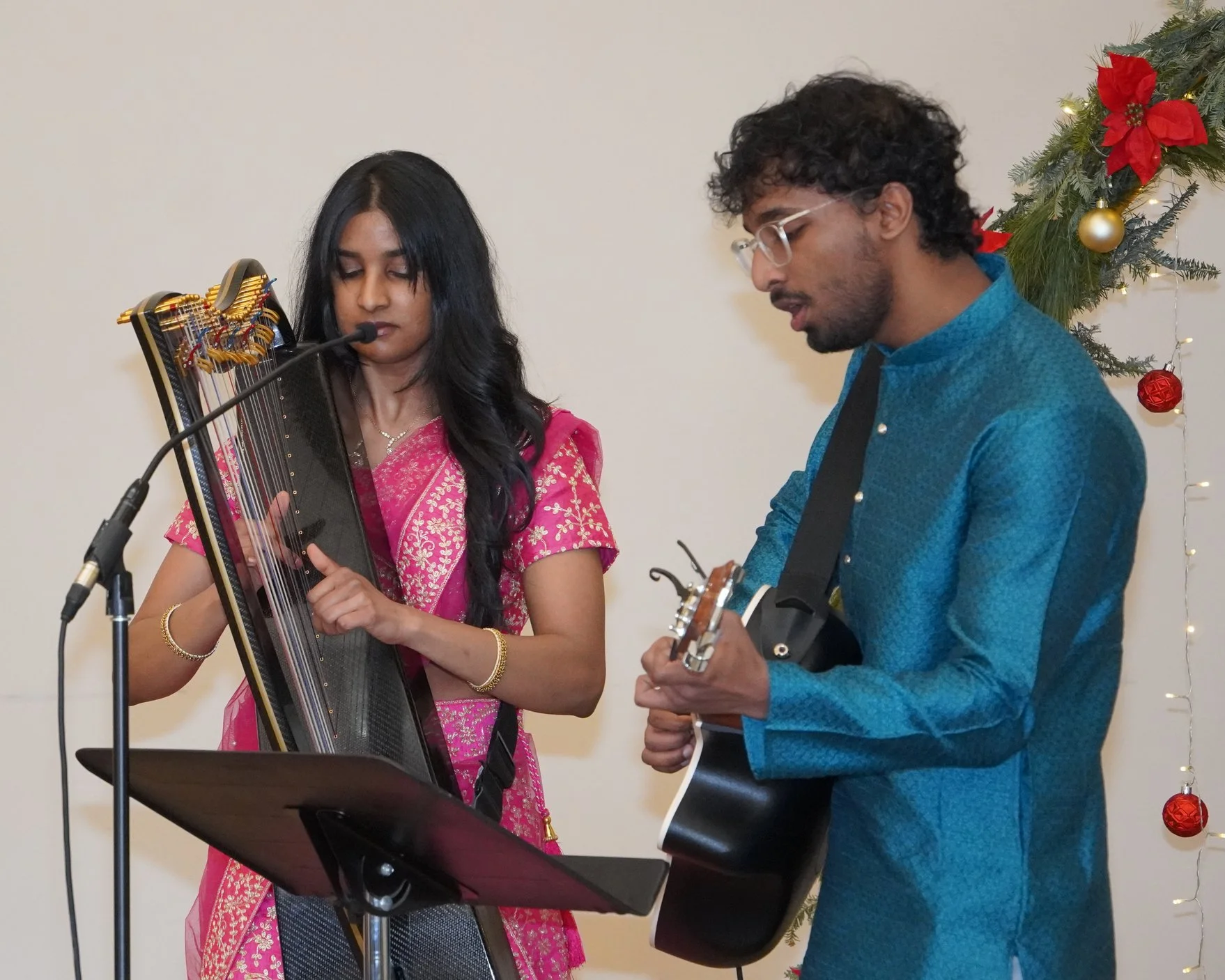 Two musicians performing in front of a Christmas tree: a woman playing a veena and a man playing an acoustic guitar.