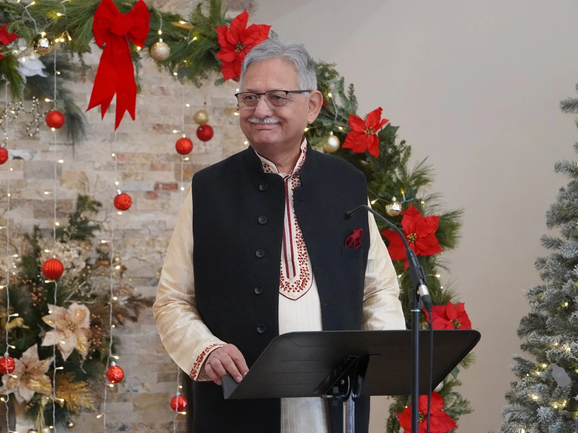 A man dressed in traditional Indian attire, standing in front of a decorated Christmas backdrop with pine branches, red bows, and ornaments, smiling at a podium.