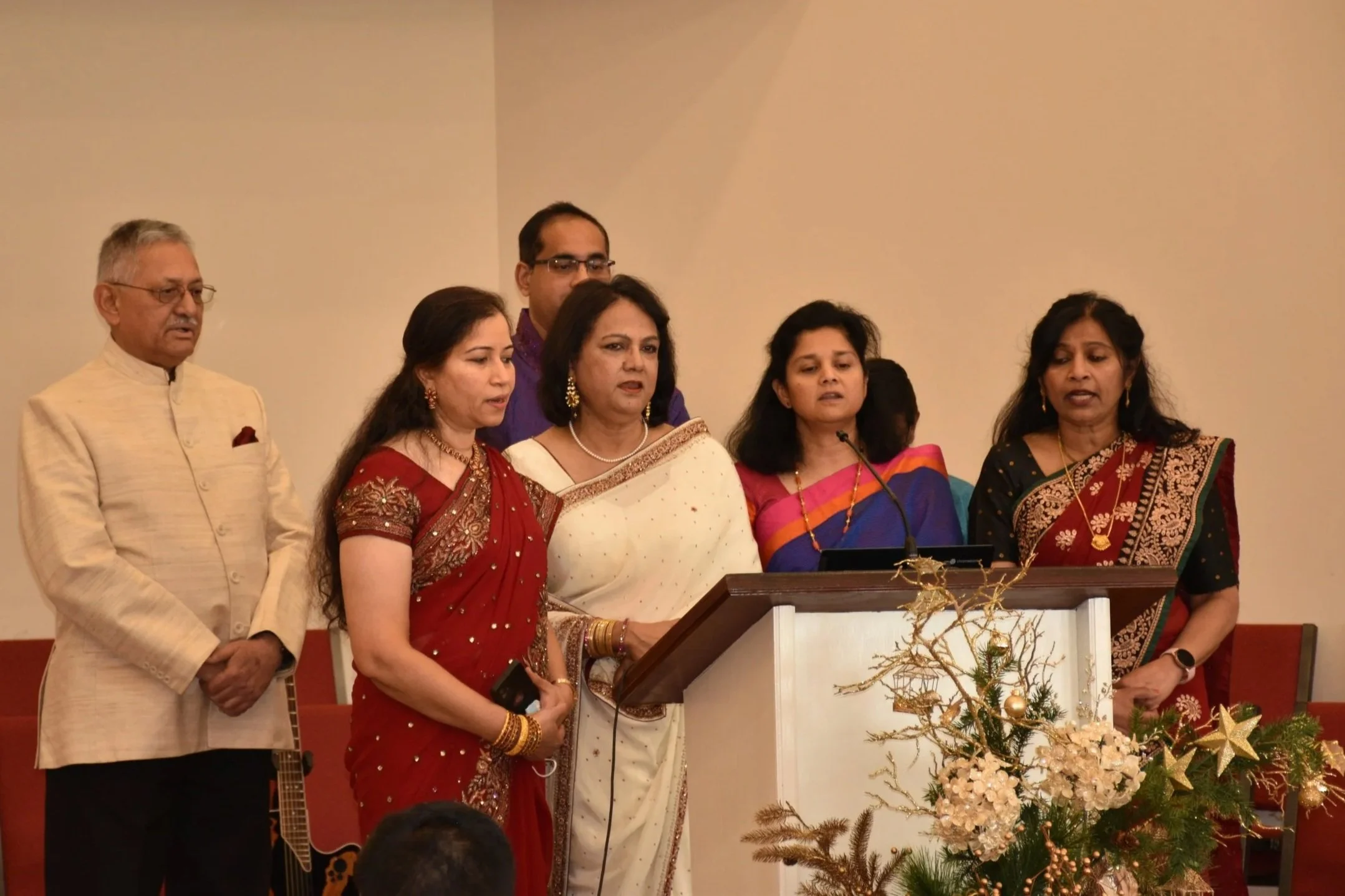 Six people standing behind a podium with Christmas decorations, dressed in traditional Indian attire, in an indoor setting.